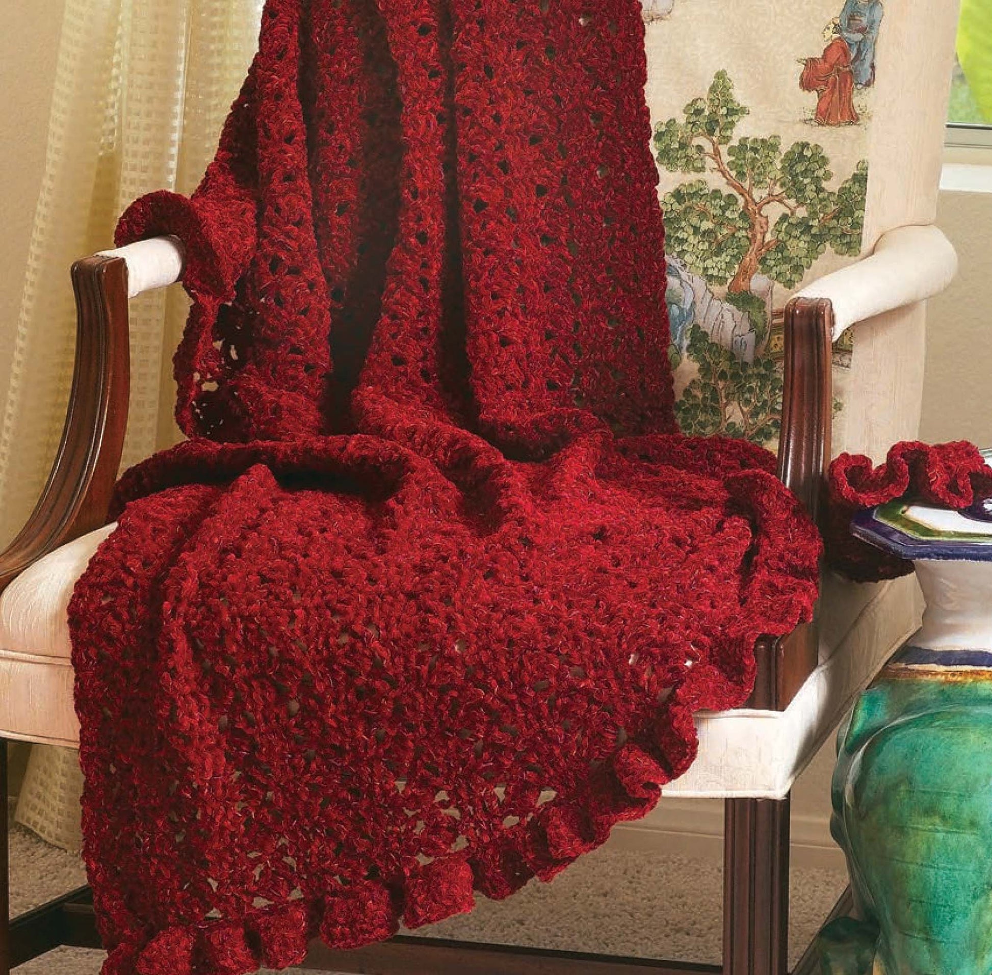 The 48-Hour Afghans Digital Download by Leisure Arts features a deep red crochet blanket with a ruffled edge, perfect for beginners, draped over a chair with a pillow and next to a ceramic table.