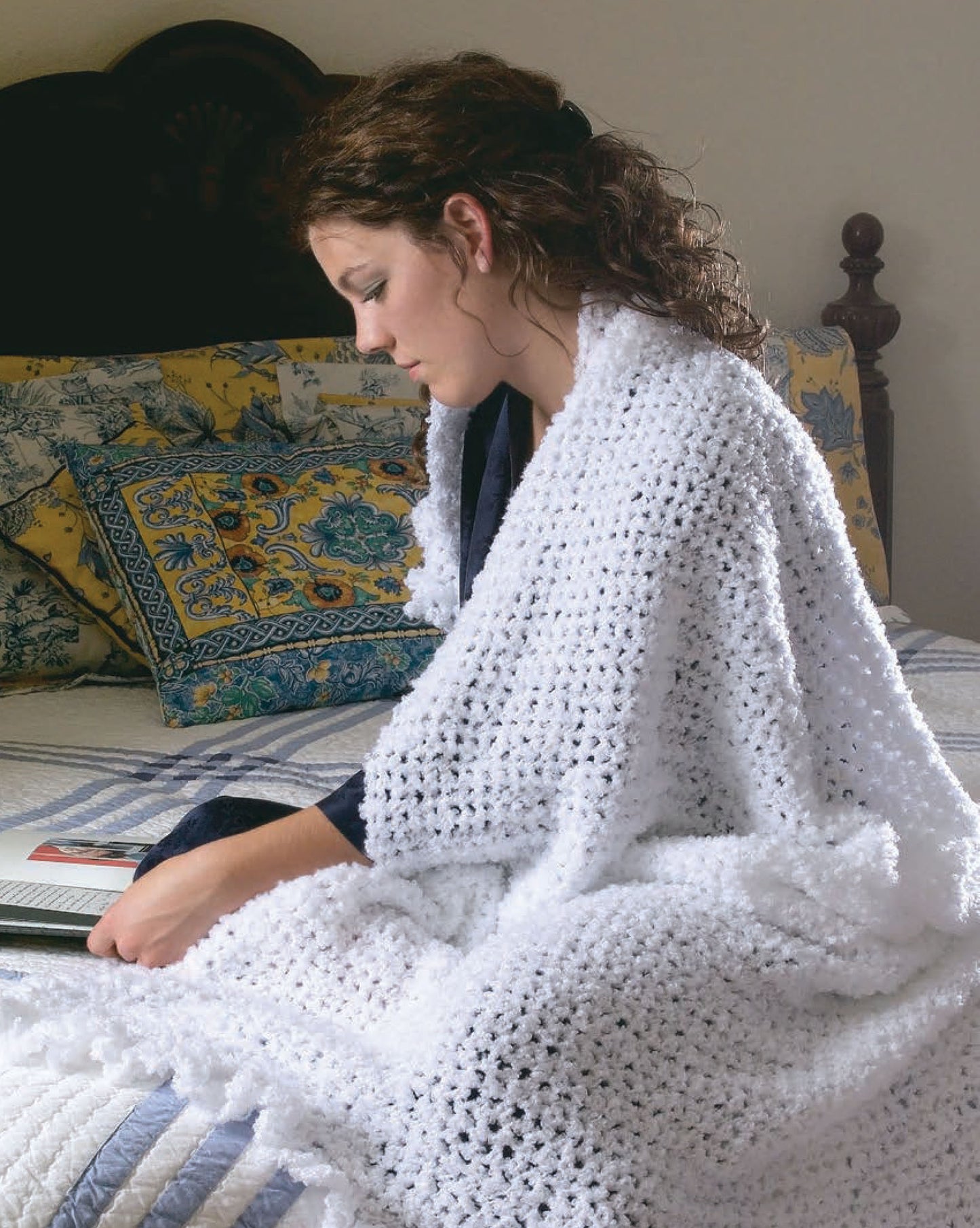 A woman with curly brown hair sits on a bed with floral pillows and a patterned quilt, wrapped in a white blanket made from Leisure Arts' 48-Hour Afghans Digital Download, reading a book.