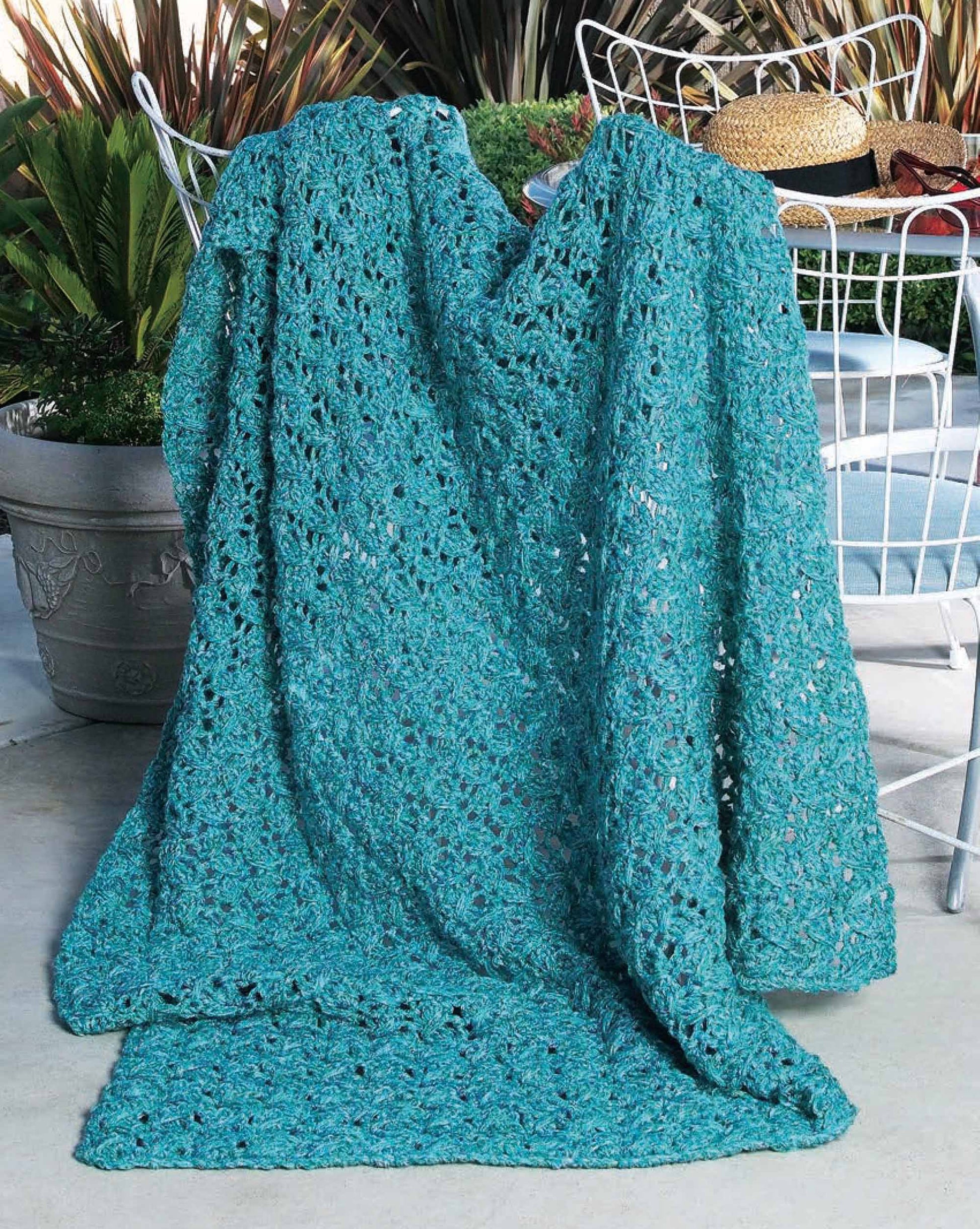 A teal afghan from Leisure Arts’ "48-Hour Afghans Digital Download" is draped over a white wire chair on a patio with potted plants—ideal inspiration for beginner crocheters.