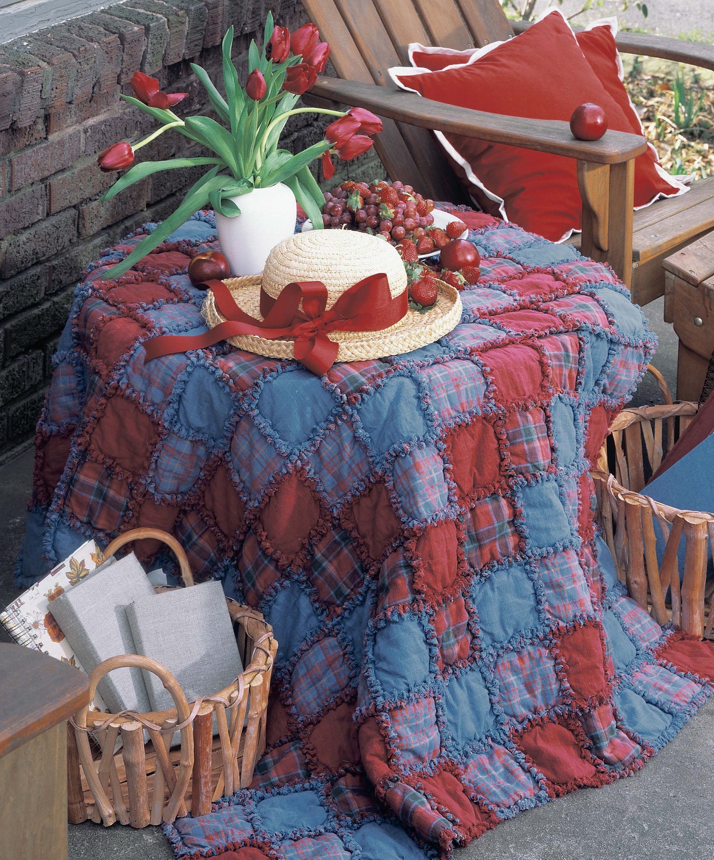 A cozy porch scene features a small table with a red-and-blue Made from Scratch Biscuit Quilts (Leisure Arts) quilt, ideal for beginners or rag quilt inspiration. On the table: a straw hat, red tulips, fruit tray. Nearby: wooden chairs and linen baskets.