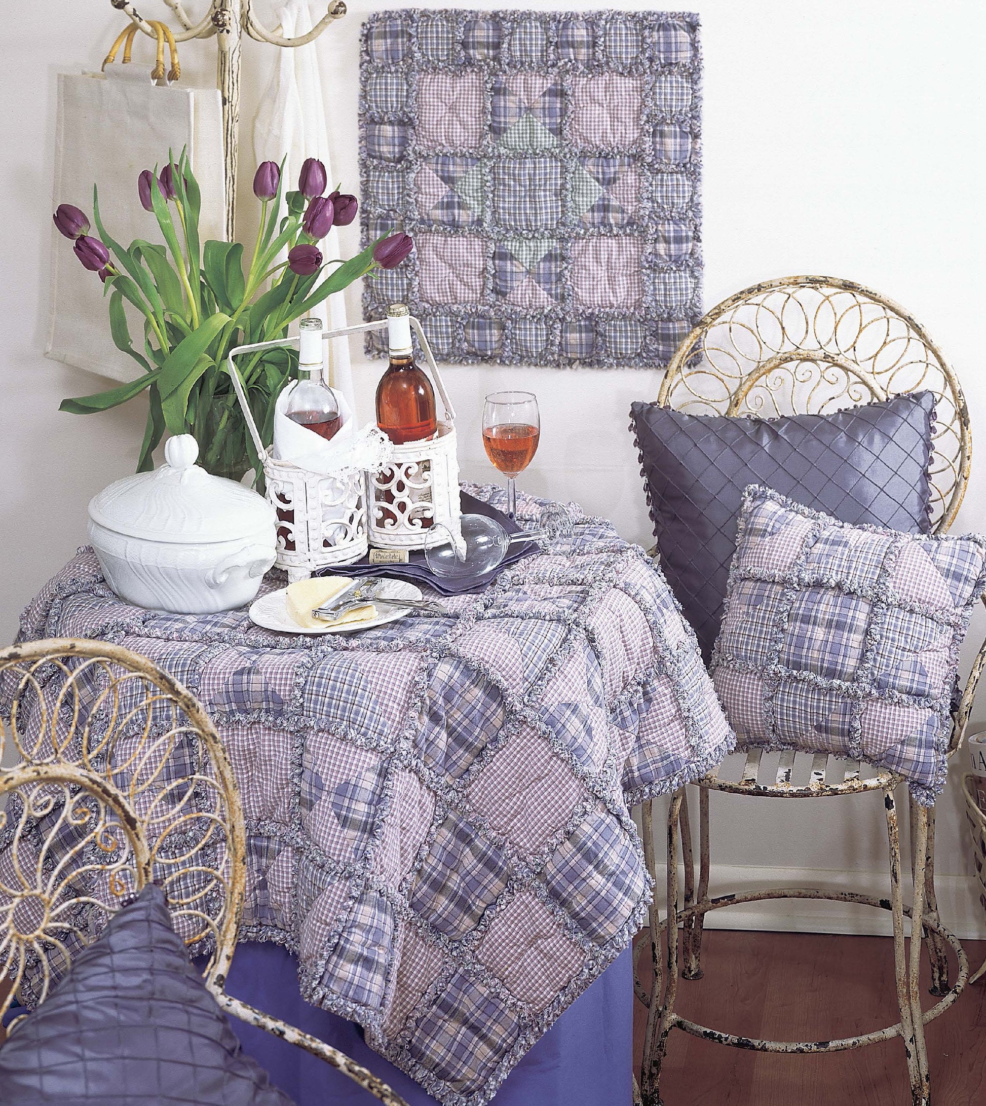 A cozy room features a small round table with a quilted purple and blue cloth inspired by Leisure Arts’ Made from Scratch Biscuit Quilts Digital Download, plus wine, glasses, tulips, and vintage chairs with matching patchwork pillows.