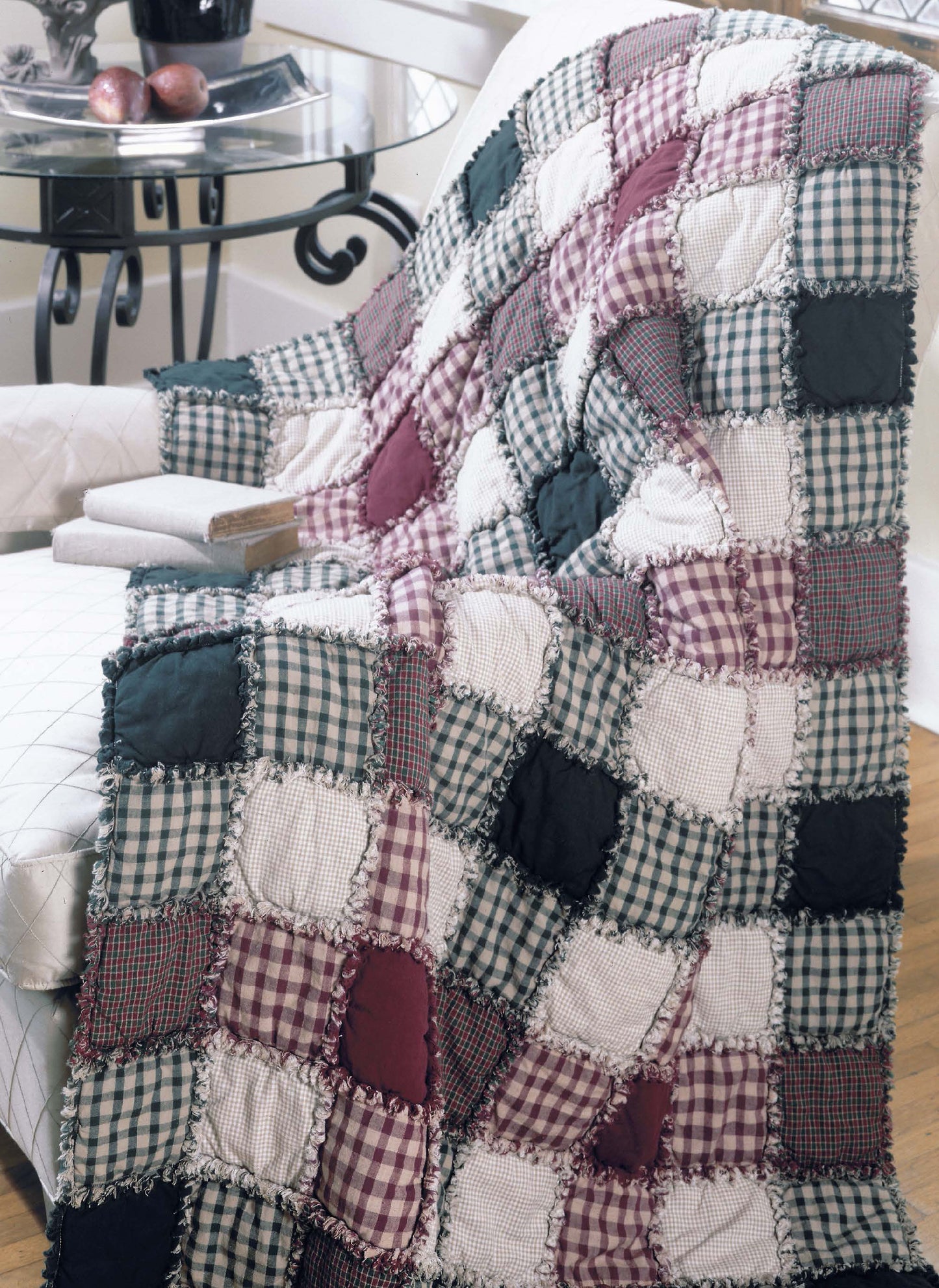The Made from Scratch Biscuit Quilts Digital Download by Leisure Arts is showcased with a quilt in burgundy, green, and cream squares draped over a white armchair. A wooden side table with apples and decor sits nearby on the floor.