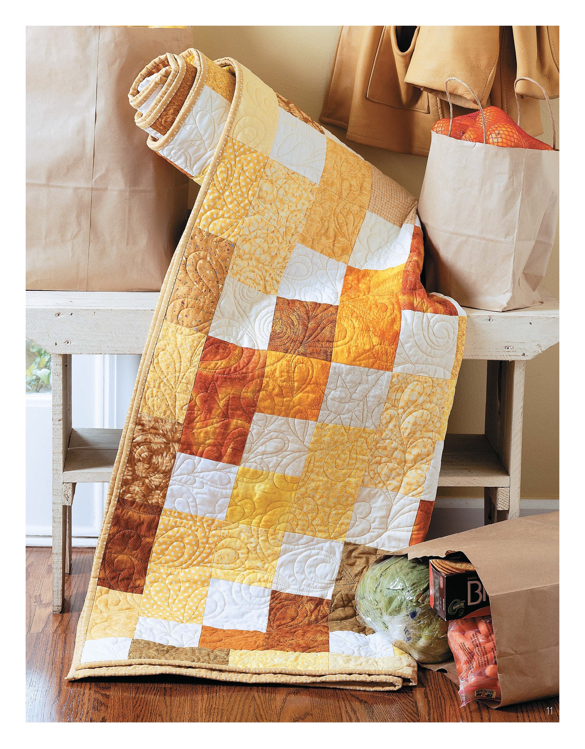 A vibrant patchwork quilt made with fast piecing from Leisure Arts’ Slash Your Stash Quilts Digital Download is draped over a bench near produce-filled grocery bags, set on a wooden floor by a cream-colored wall.