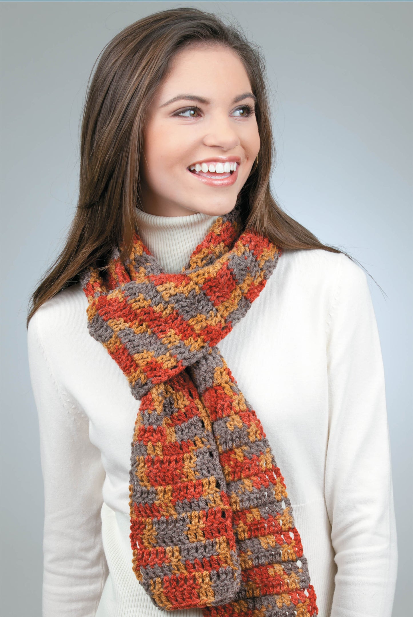 A woman wearing a cream turtleneck and a colorful crocheted scarf—made with Leisure Arts’ "Learn to Crochet Now!" digital download—smiles and looks left against a plain gray background.