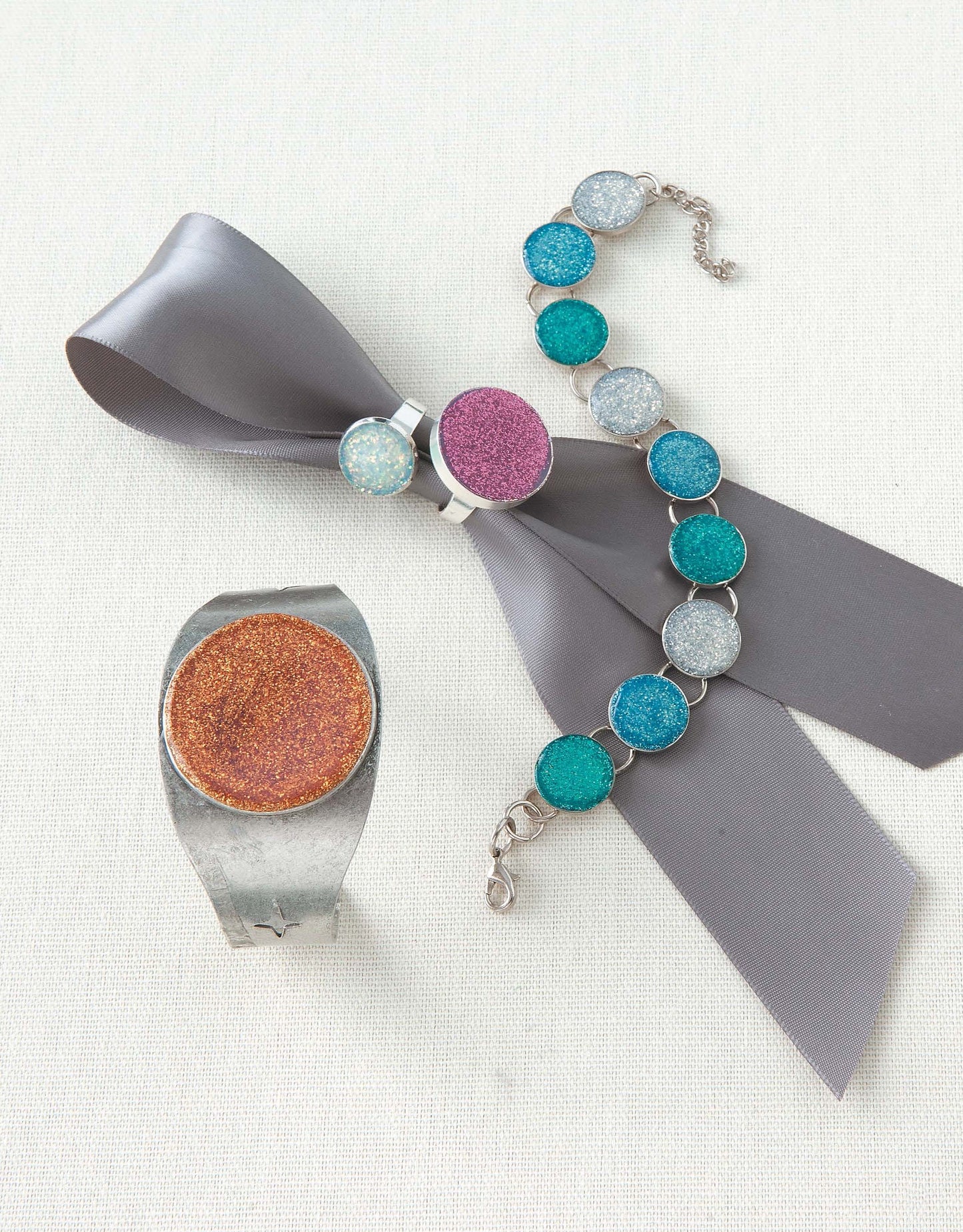 The Oh-So-Easy Jewelry Digital Download by Leisure Arts is ideal for beginners, featuring a silver ring with a glittery orange stone, a gray ribbon with a purple brooch, and a bracelet adorned with blue and silver glittery stones.