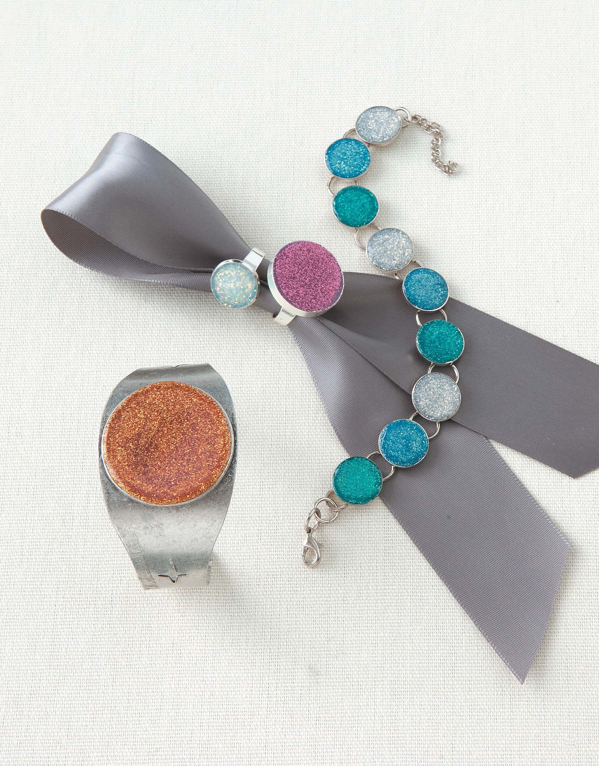 The Oh-So-Easy Jewelry Digital Download by Leisure Arts is ideal for beginners, featuring a silver ring with a glittery orange stone, a gray ribbon with a purple brooch, and a bracelet adorned with blue and silver glittery stones.