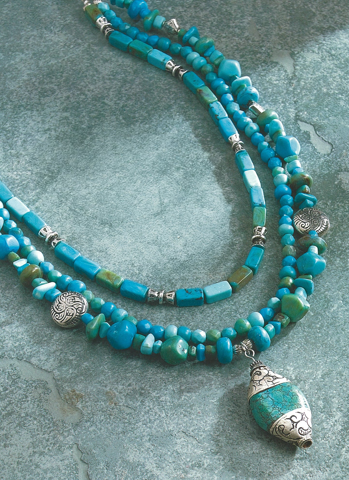 The Leisure Arts Pendant Necklaces feature three turquoise blue beaded strands with silver accents. The longest strand displays a large pendant with turquoise stones and silver detailing, showcased on a textured gray surface.