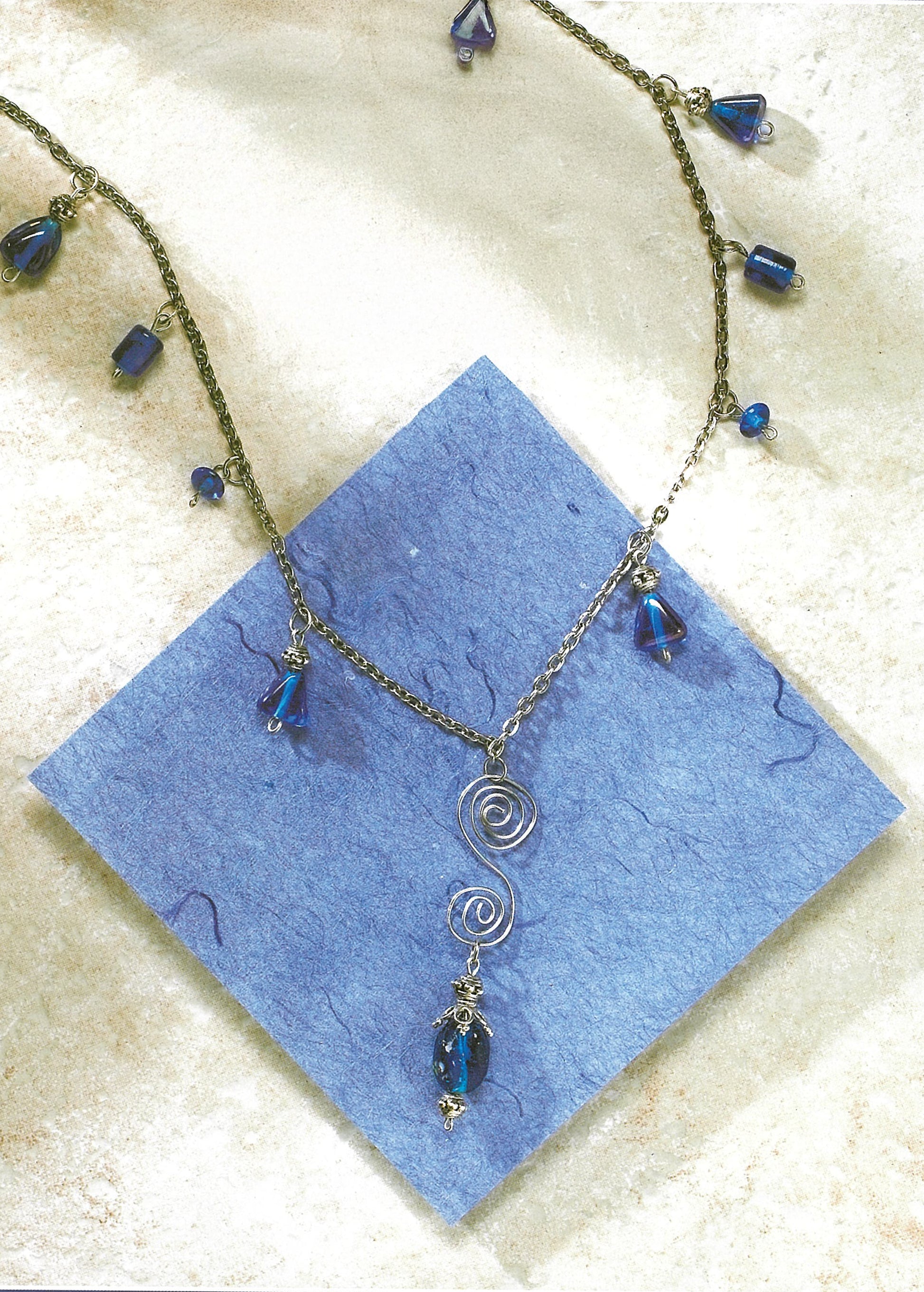 The Leisure Arts Pendant Necklaces feature blue glass beads and a wire spiral pendant, ideal for beaded jewelry lovers, displayed on textured blue paper atop a light stone surface.