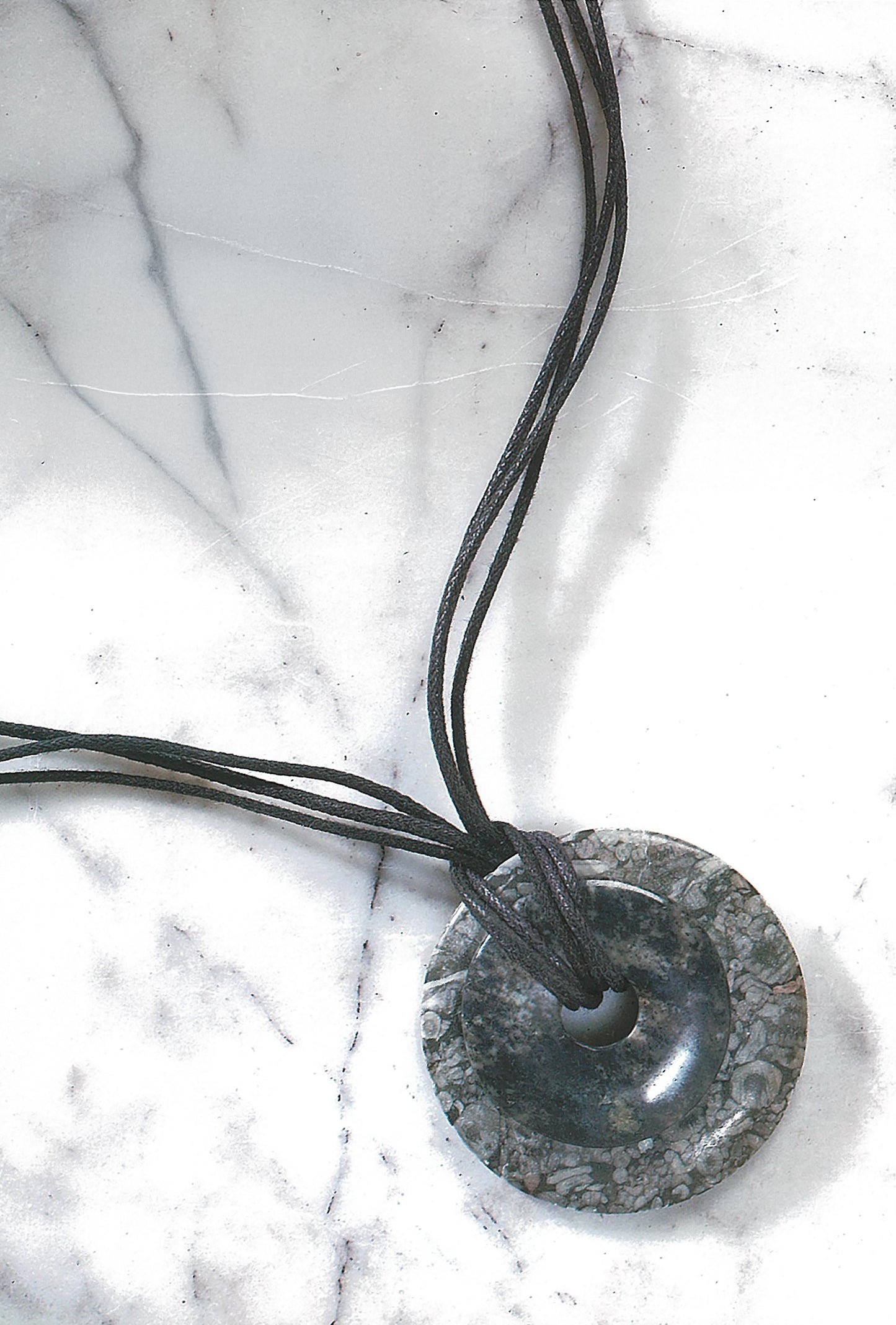 The Pendant Necklaces by Leisure Arts feature a round, dark polymer clay pendant with a textured pattern, tied with black cords and displayed on white marble with grey veins.