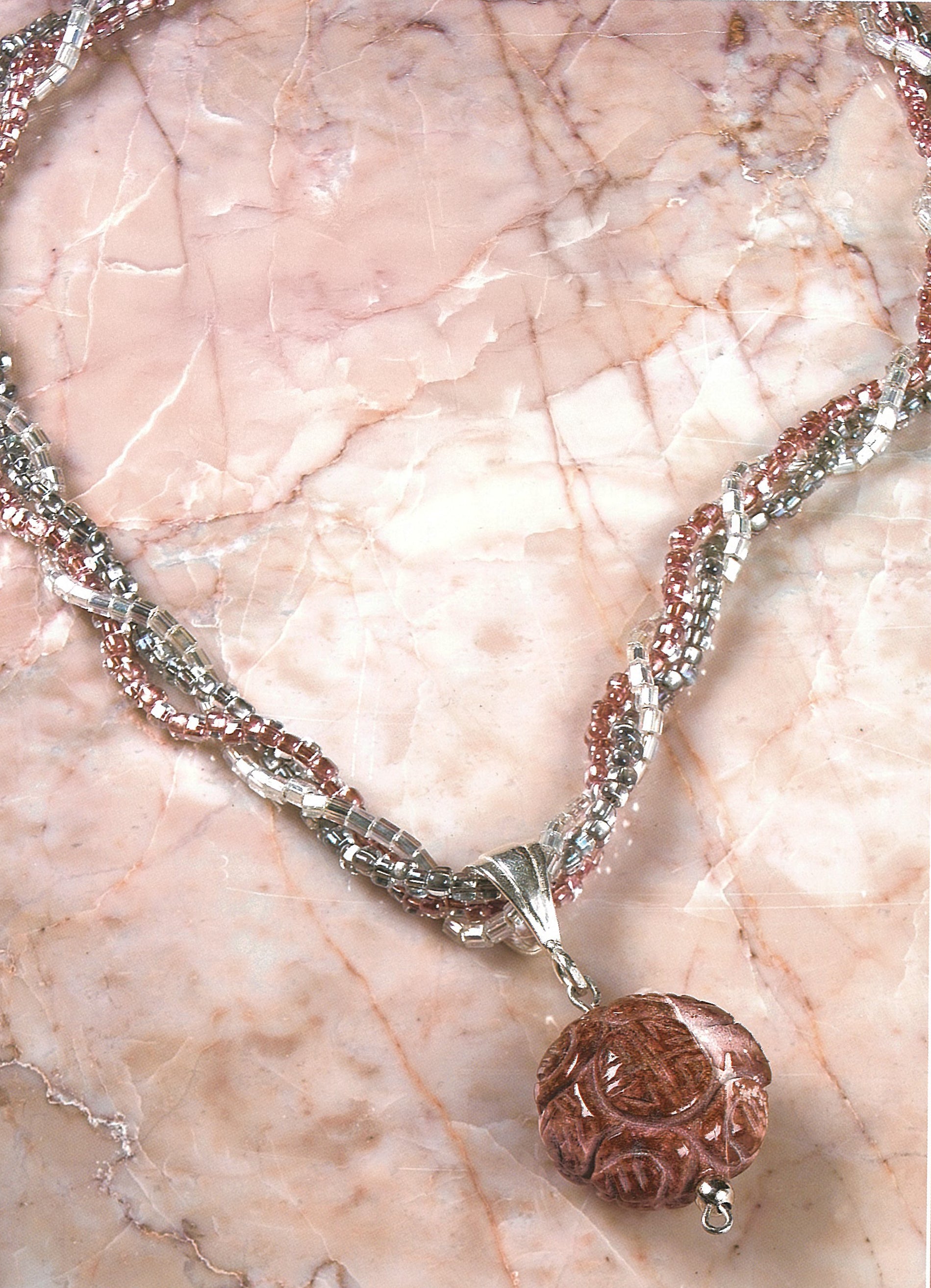 The Pendant Necklaces by Leisure Arts feature twisted pink and silver beads with a carved polymer clay pendant, beautifully displayed on polished pink marble.