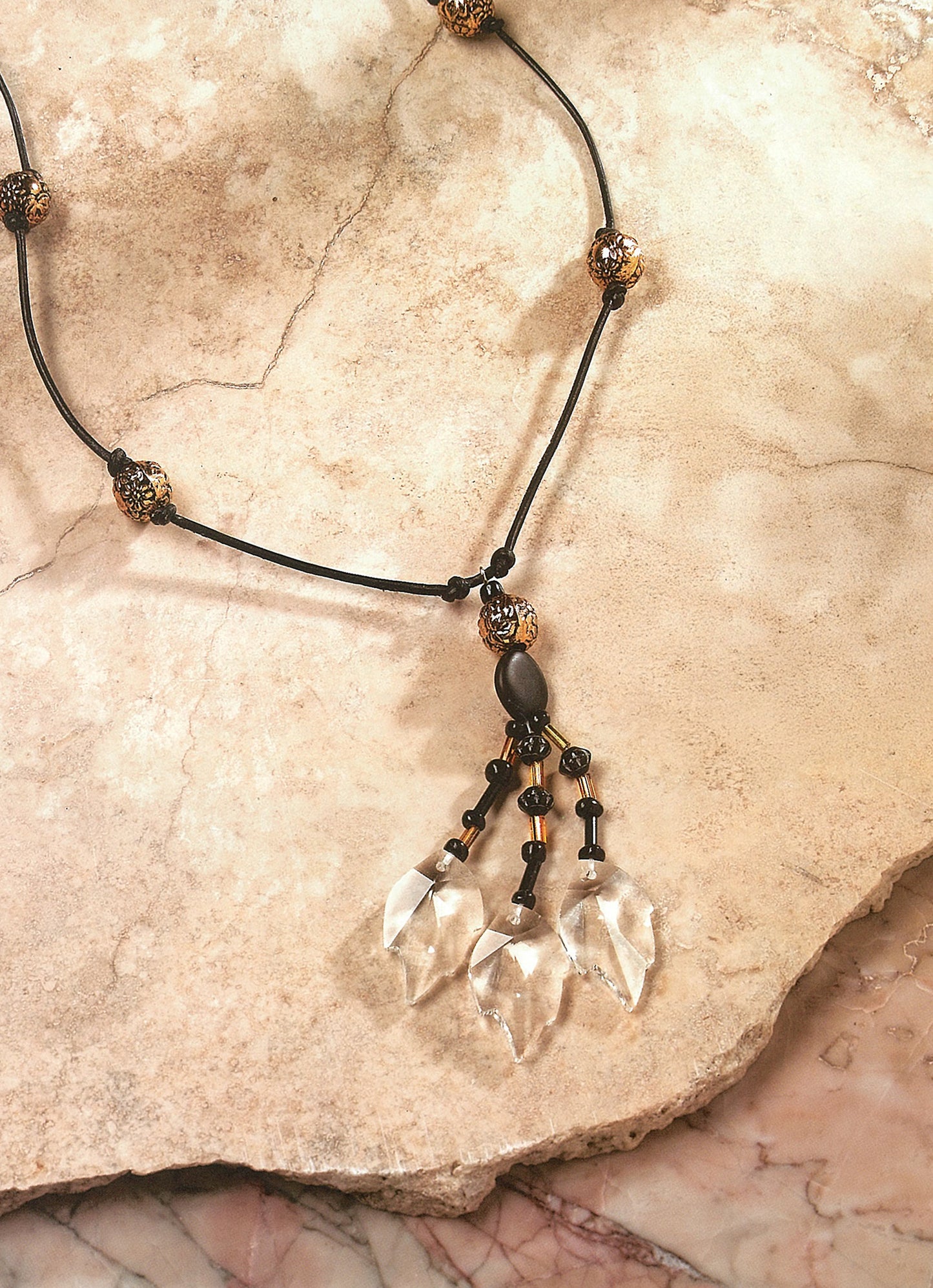 The Pendant Necklaces by Leisure Arts feature a black cord, metallic round beads, and a pendant with three clear crystal-like dangles, shown on a beige and pink marbled stone background.