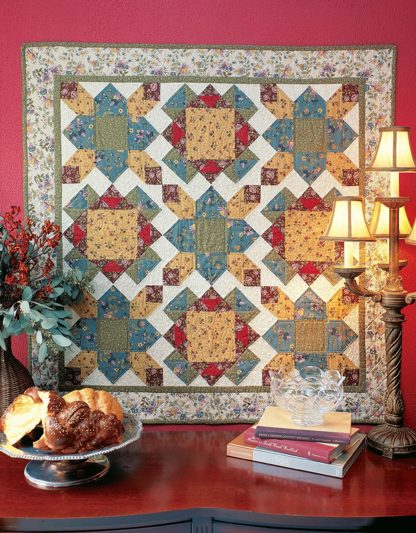 A vibrant patchwork quilt made from the Quilter's Mix & Match Blocks by Leisure Arts, with blue, yellow, red, and cream patterns, hangs above a wooden table holding books, crystal dishes, a lamp, fresh flowers in a vase, and braided bread.