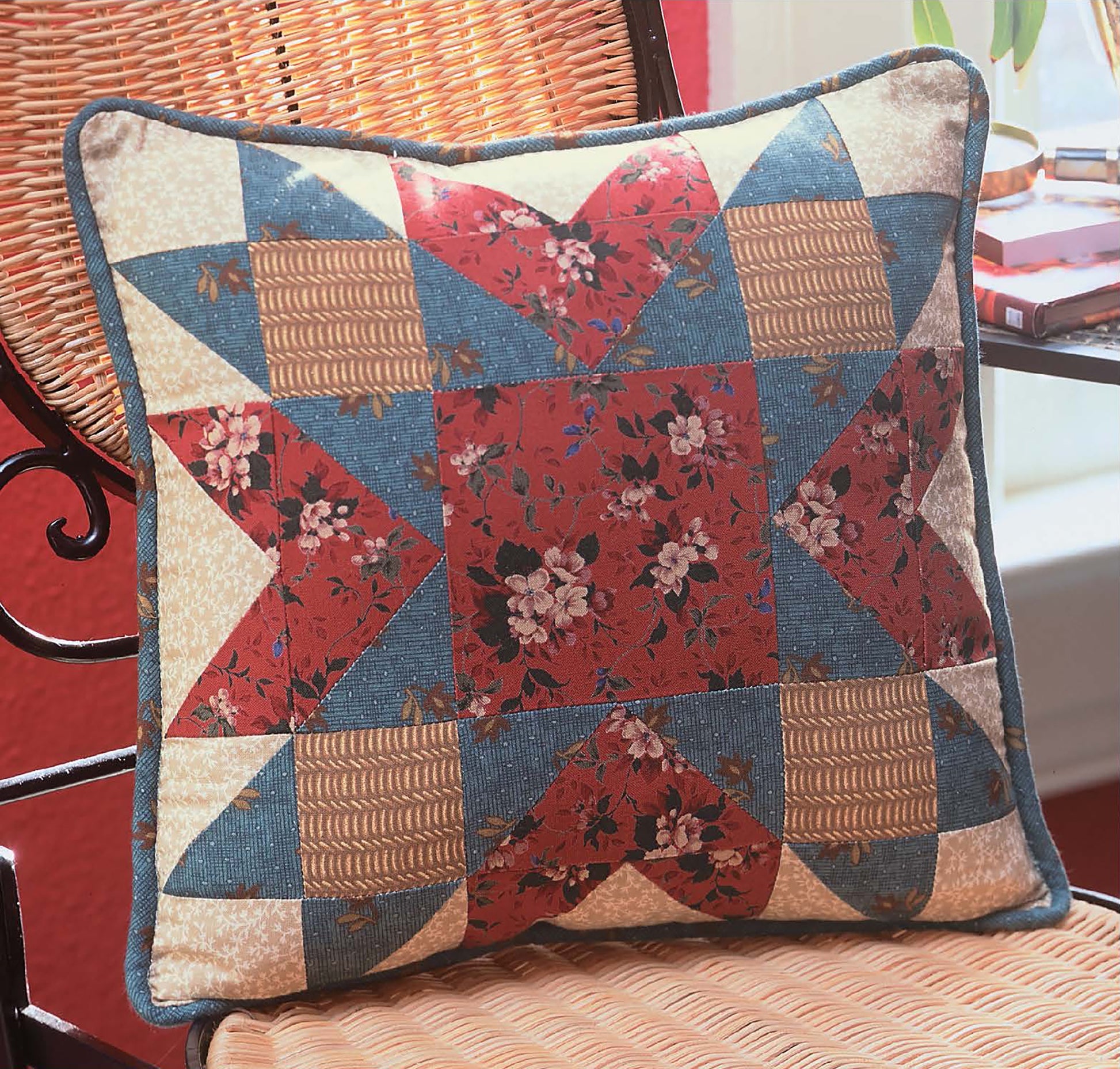 The Quilter's Mix & Match Blocks throw pillow by Leisure Arts features a vibrant patchwork of red, blue, tan, and floral fabrics—perfect inspiration for quilting projects. It’s shown on a wicker chair by the window.