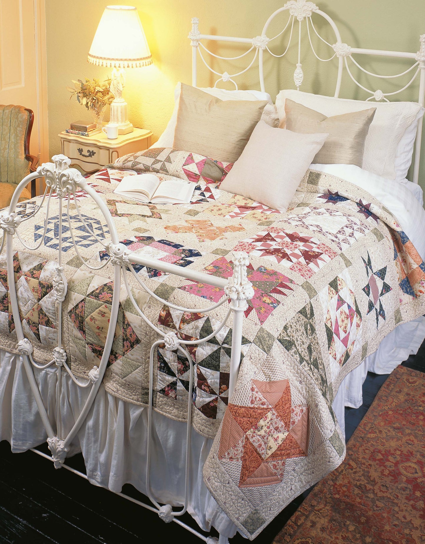 A cozy bed features Leisure Arts' Quilter's Mix & Match Blocks quilt, vibrant pillows, and an open book—all in a warmly lit bedroom with a vintage lamp, nightstand, and decorative metal headboard and footboard.