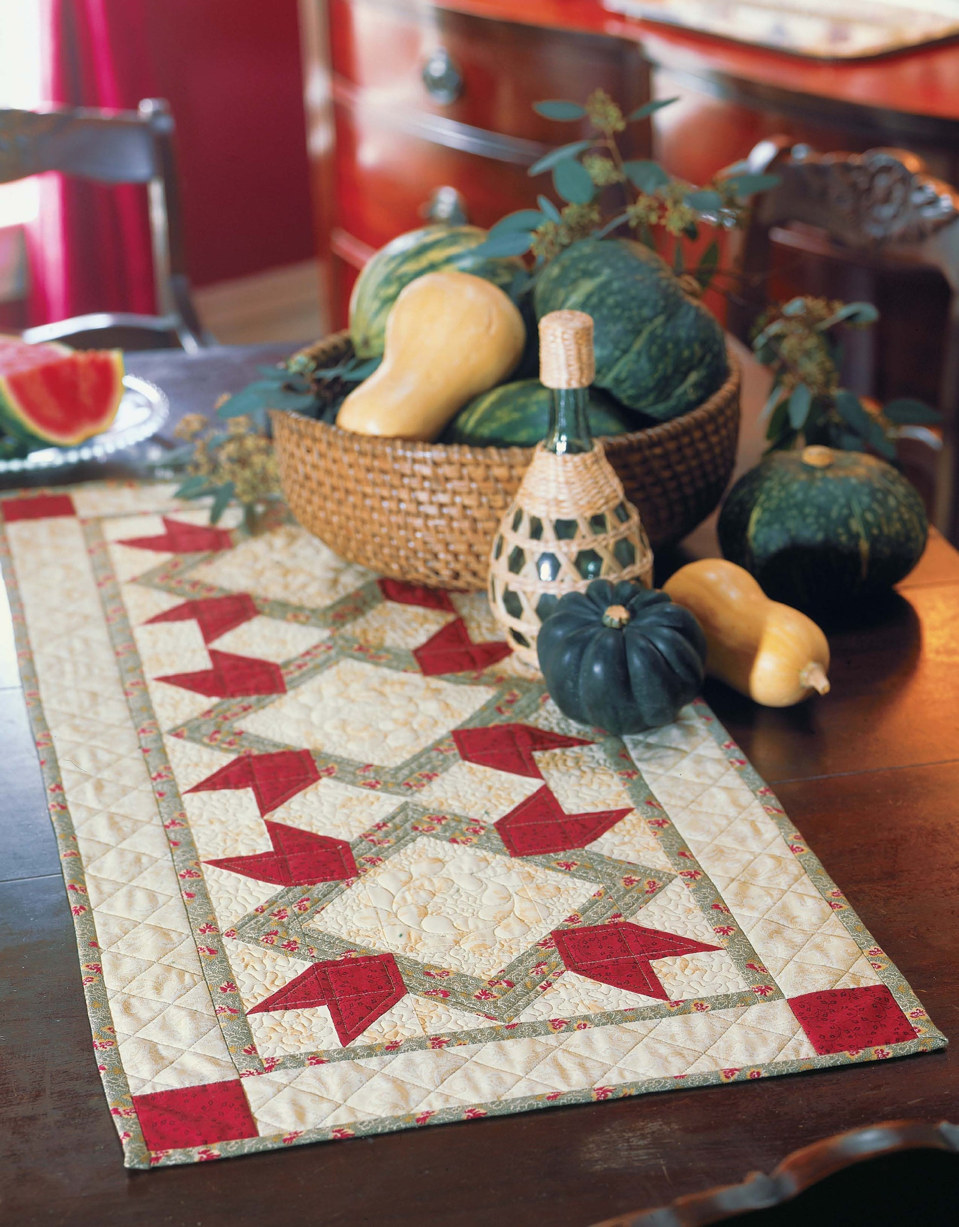 A woven basket of squashes and gourds and a bottle sit on a dark wooden table with a decorative quilted runner—perfect inspiration for your next project with Leisure Arts' Quilter's Mix & Match Blocks. A sliced watermelon appears in the background.