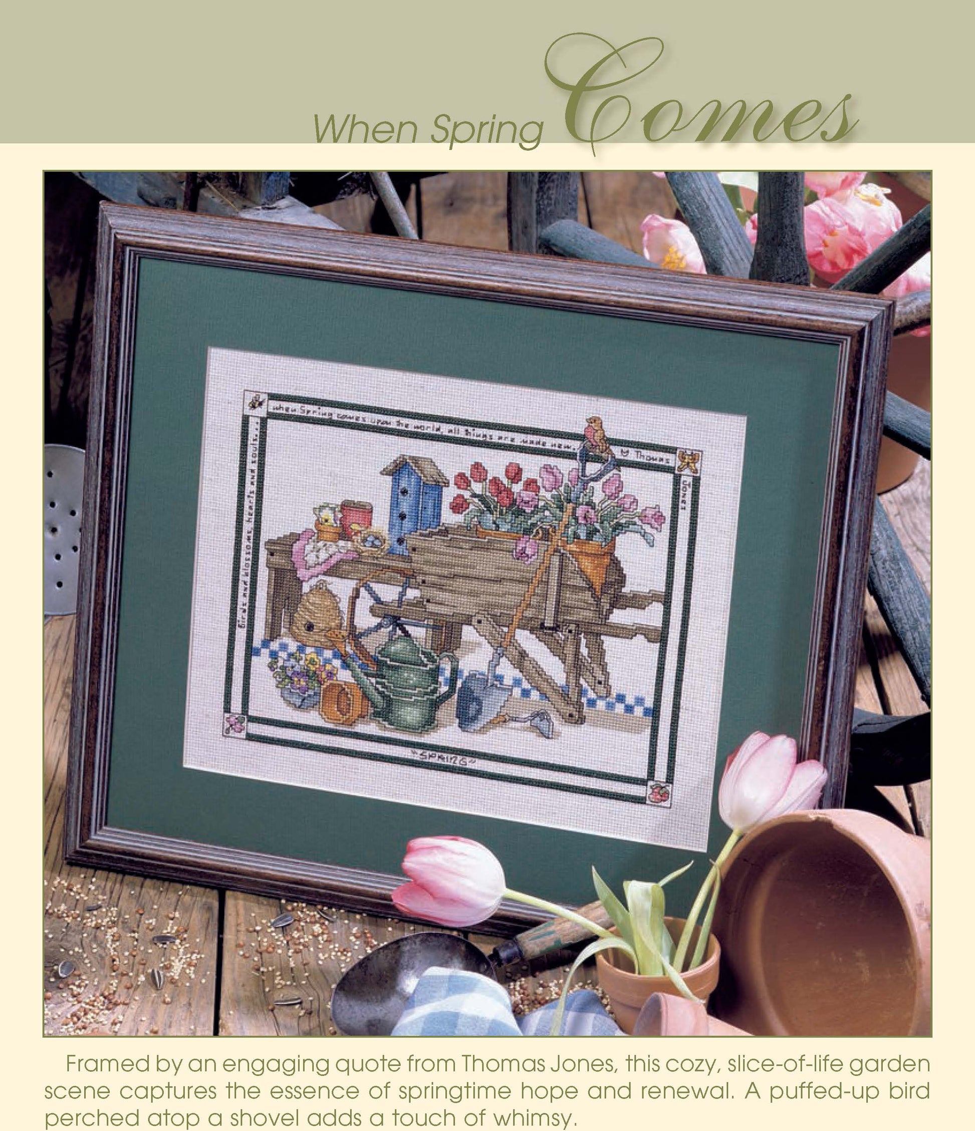 The Simply Inspirational cross stitch by Leisure Arts features a spring garden with a wheelbarrow, flowers, birdhouse, watering can, and bird on shovel. Displayed framed on a wooden table with scattered tulips and garden tools.