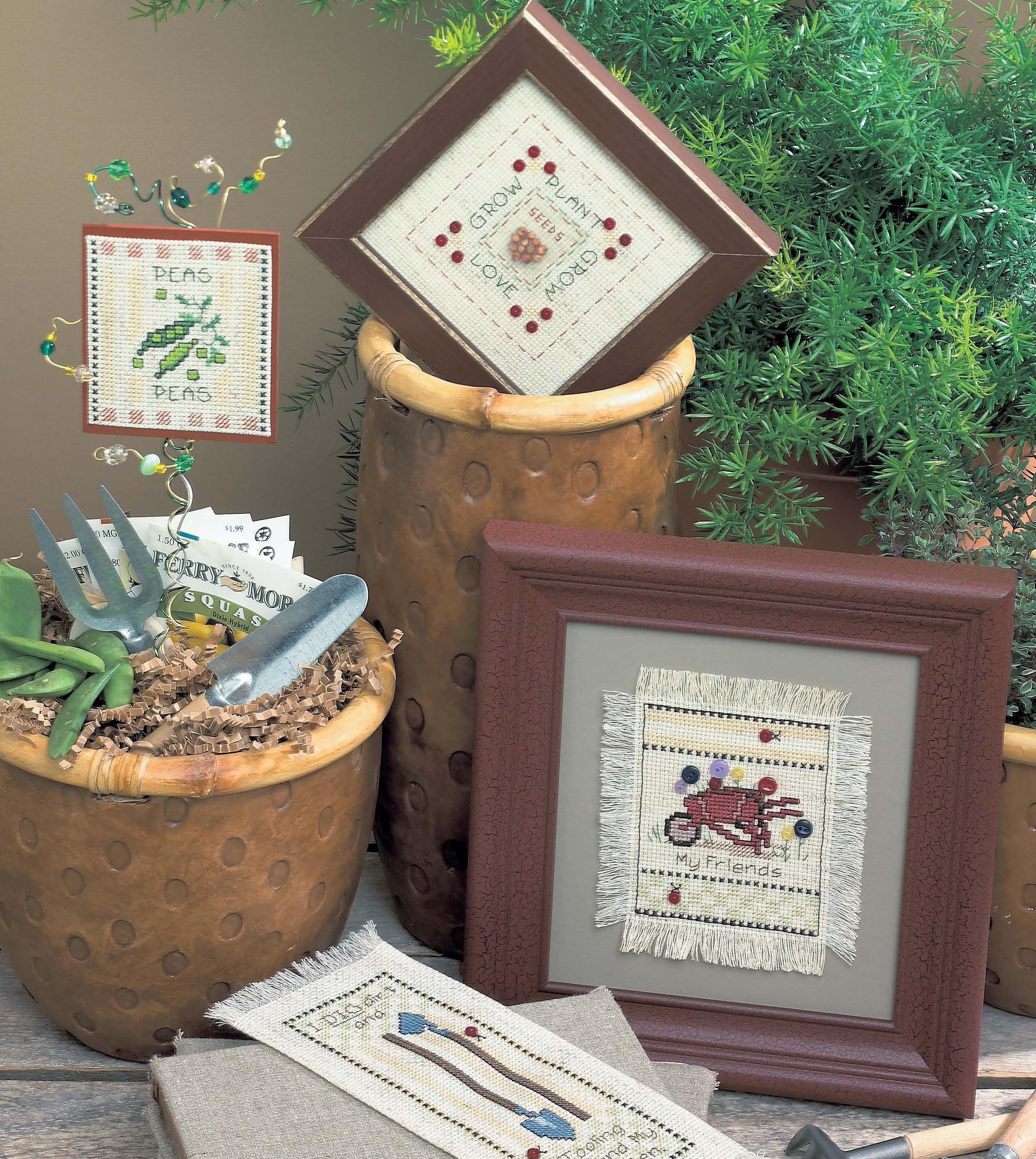 Framed "Life Tips from the Garden" cross stitch by Leisure Arts, with garden and friend themes, is displayed among brown pots, Aida cloth, gardening tools, and green plants for a cozy rustic touch.
