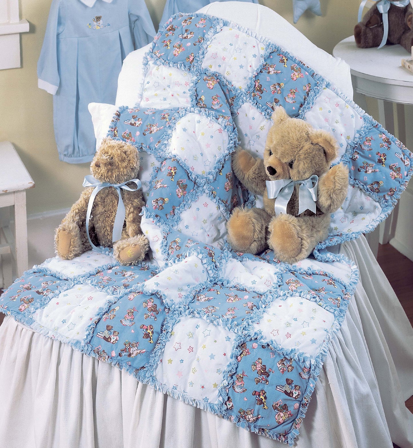 Two teddy bears with blue bows sit on a white bench beside a patchwork quilt featuring playful bears. In the background, a blue shirt hangs. Create your own with "Made from Scratch Biscuit Quilts" by Leisure Arts—ideal for baby and patchwork projects!.