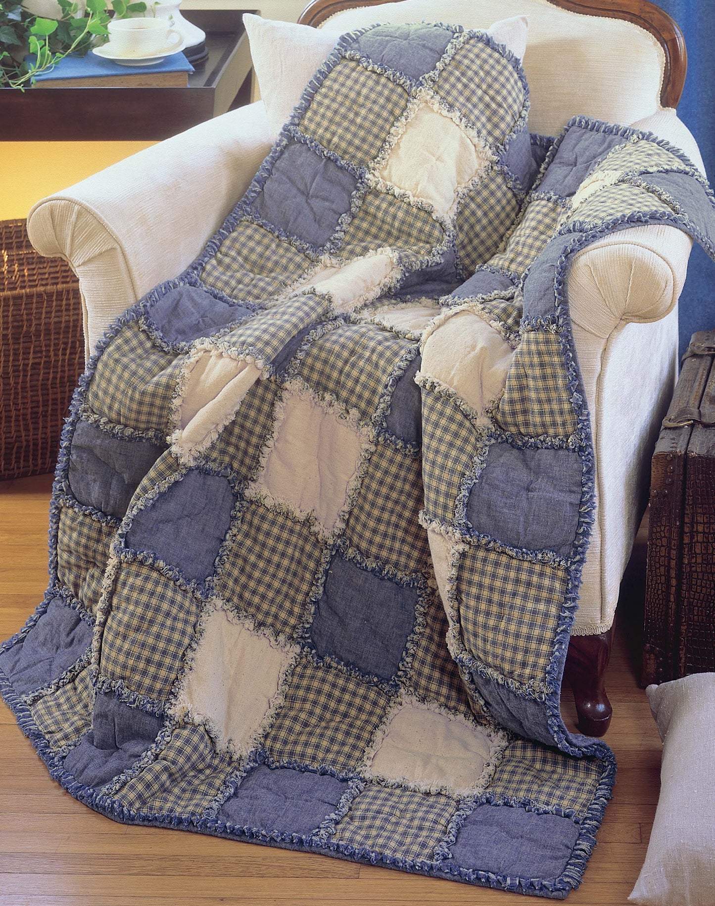 The Made from Scratch Biscuit Quilts by Leisure Arts is a cozy patchwork quilt in blue, beige, and checkered patterns, draped over an armchair beside a wooden table with a teacup and rustic trunk on a wooden floor.