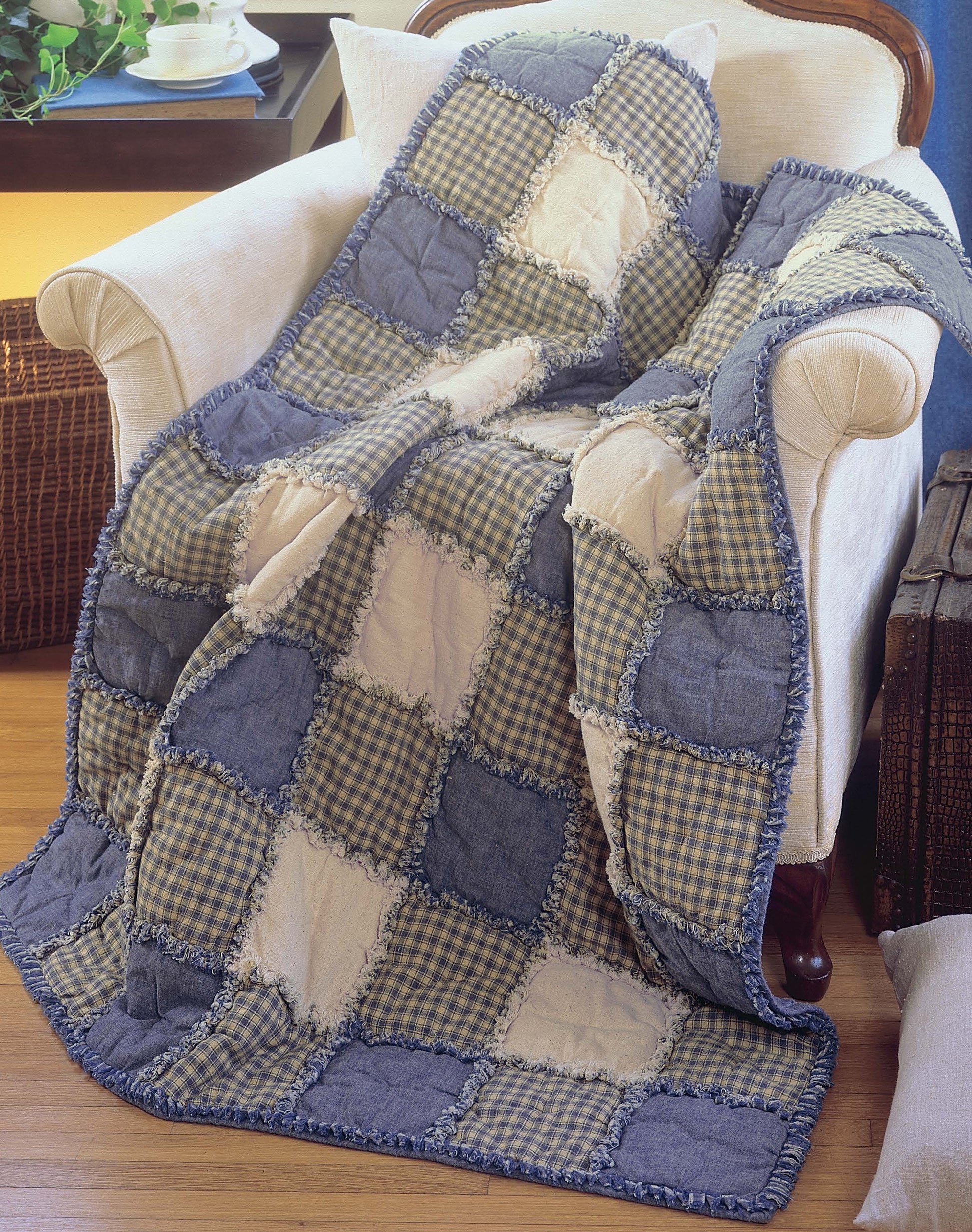 The Made from Scratch Biscuit Quilts by Leisure Arts is a cozy patchwork quilt in blue, beige, and checkered patterns, draped over an armchair beside a wooden table with a teacup and rustic trunk on a wooden floor.