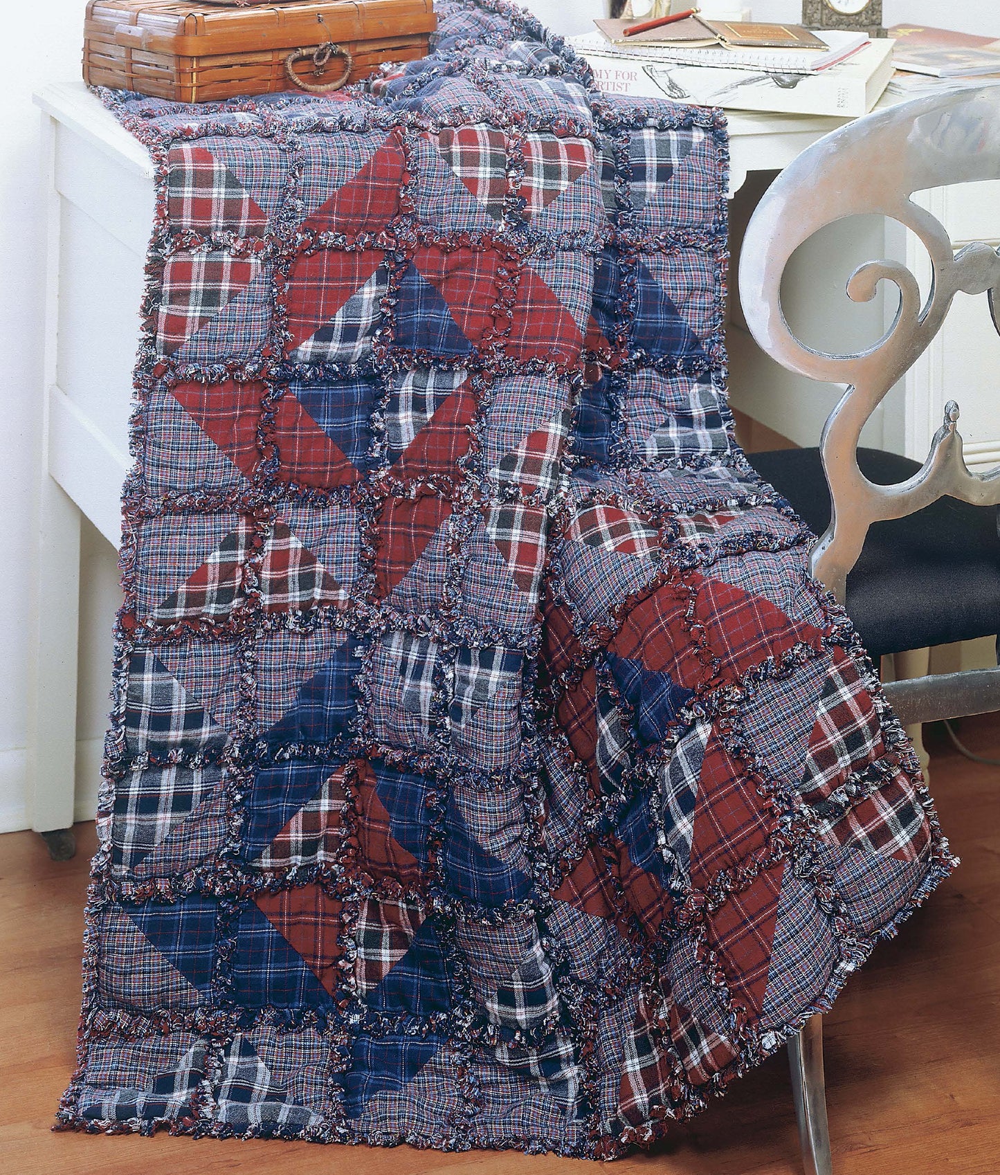 The Made from Scratch Biscuit Quilts by Leisure Arts, featuring red, blue, and plaid fabric squares, is perfect for patchwork or rag quilt projects—shown draped over a white desk with a silver chair and wooden box amidst books.
