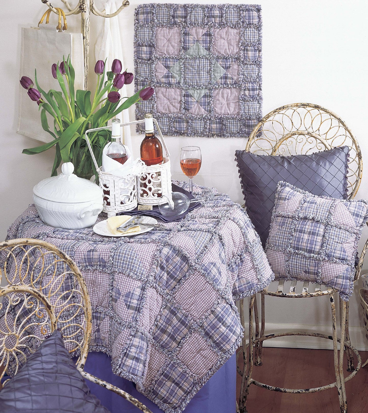 A cozy setting features a small round table with wine, glasses, and plates, surrounded by wicker chairs. Purple tulips and the Made from Scratch Biscuit Quilts by Leisure Arts add homespun charm to the bouquet and wall decor.