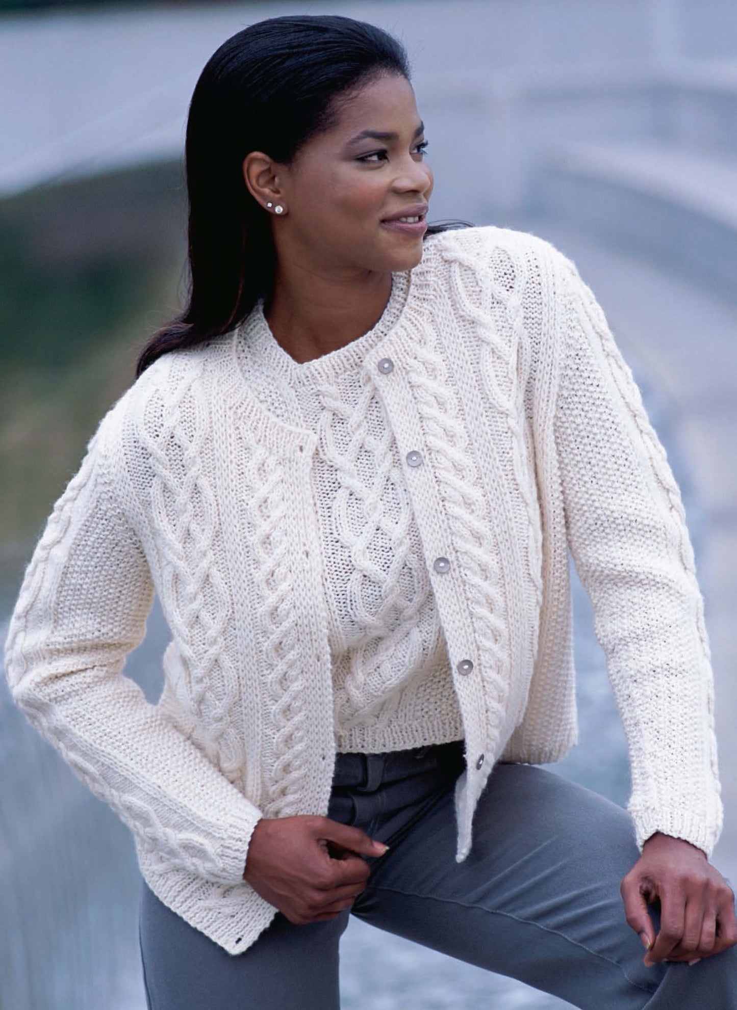 A woman models a cream cable-knit sweater and cardigan from Better Homes and Gardens Knitted Sweaters for Her by Leisure Arts, pairing them with gray pants as she smiles outdoors near a water feature.