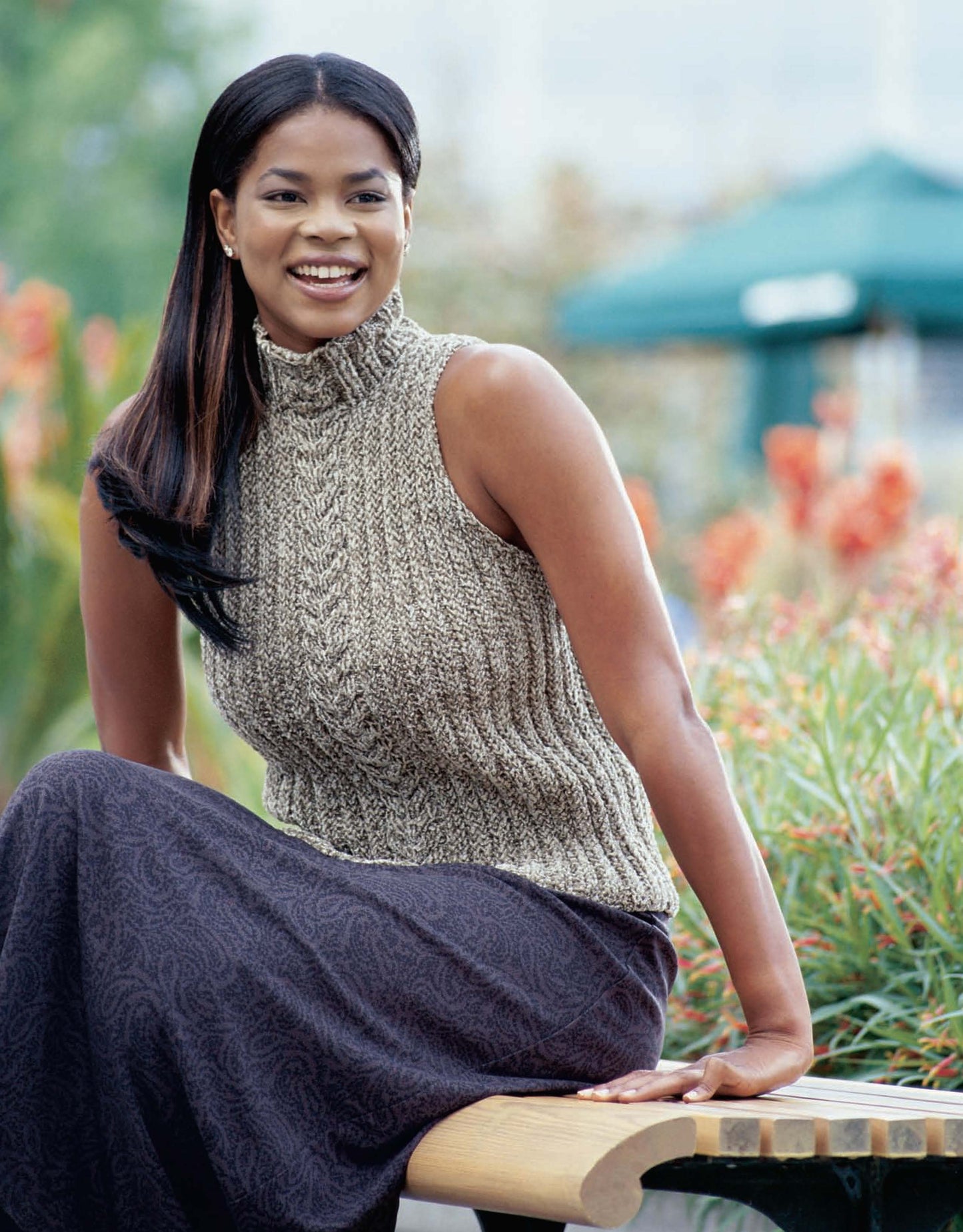 A woman with long, dark hair smiles while seated on a wooden bench outdoors, wearing a sleeveless high-neck sweater from Better Homes and Gardens Knitted Sweaters for Her by Leisure Arts, with bright orange flowers and greenery in the background.