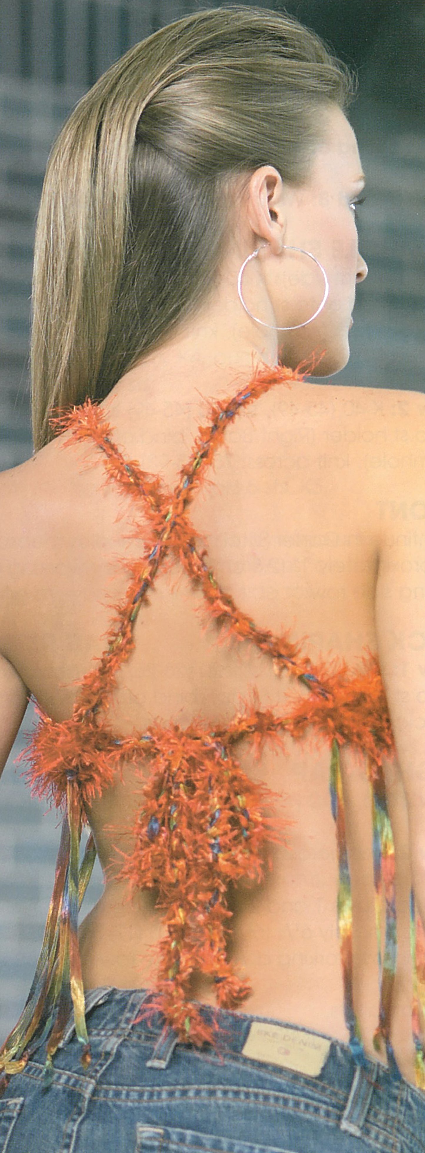 A model wears the Knit Tops for You by Leisure Arts, featuring a backless shoulder design with orange eyelash yarn crisscross straps, paired with large hoop earrings, a sleek ponytail, and low-rise jeans.