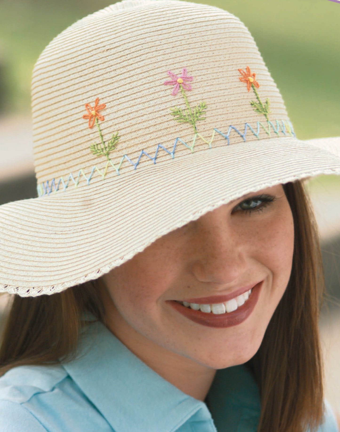 A smiling woman models the Sew Simple Embroidery wide-brimmed straw hat by Leisure Arts, featuring colorful embroidery and zigzag patterns, paired with a light blue collared shirt against a blurred green background.