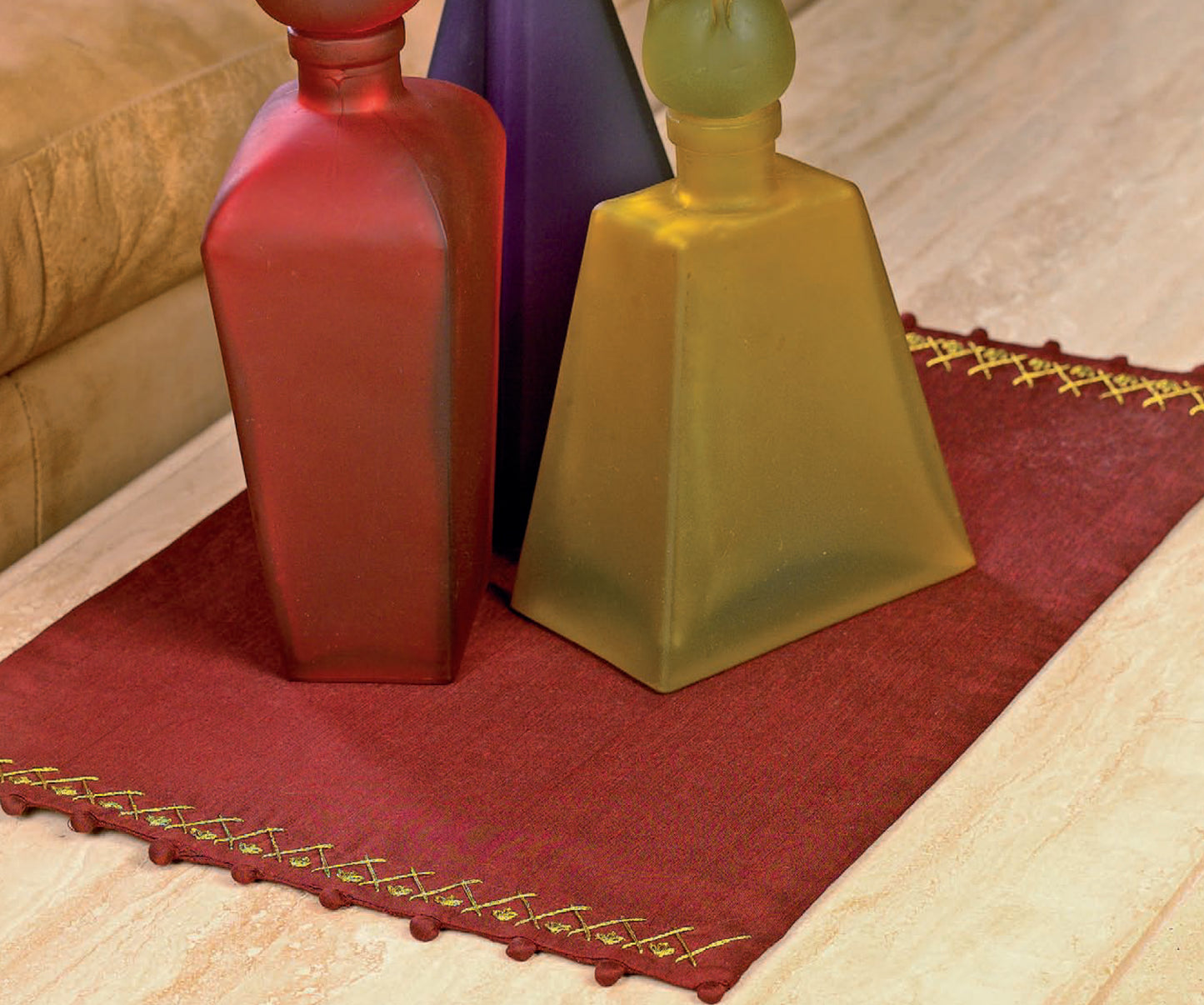 Three frosted glass bottles—red, yellow, and purple—sit on a red mat with yellow edging, displaying beginner projects from Sew Simple Embroidery by Leisure Arts and adding charm to the light marble surface.