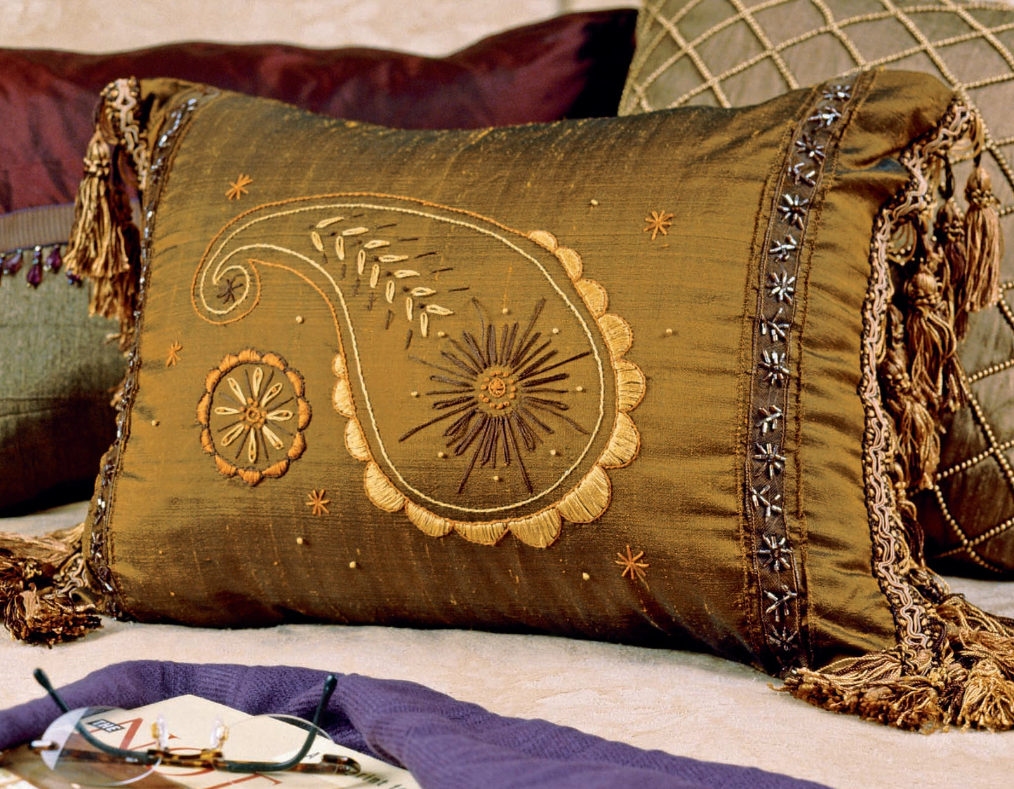 A Sew Simple Embroidery rectangular throw pillow from Leisure Arts, with gold embroidery, paisley and floral patterns, rests on a couch among other designer originals. Glasses and a purple cloth sit nearby.