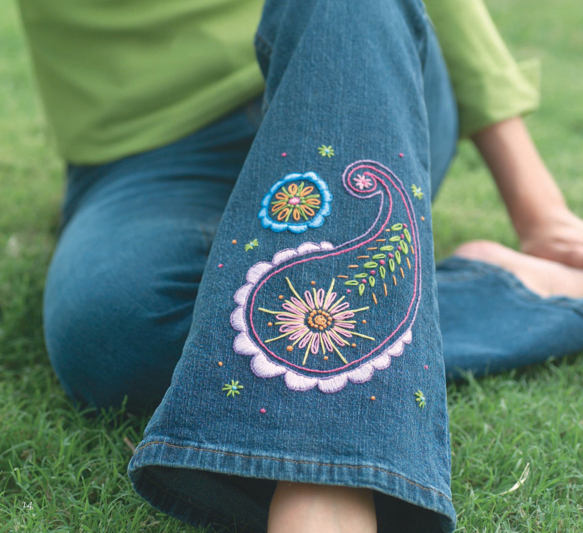 A person sits cross-legged on grass in blue jeans with colorful embroidered floral and paisley designs, a great inspiration for unique projects using Sew Simple Embroidery by Leisure Arts. They are also wearing a light green top.