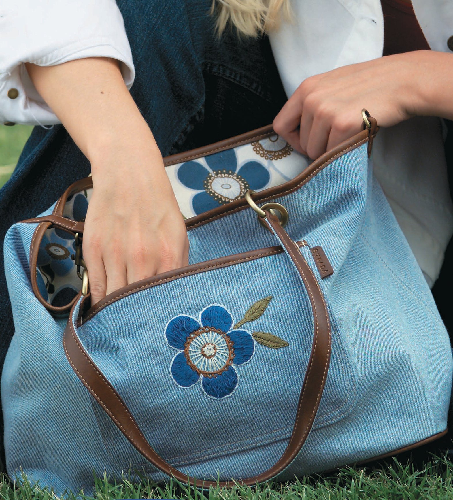 A person reaches into the Sew Simple Embroidery handbag by Leisure Arts, a blue denim bag with brown leather straps, blue flower applique, and floral lining—ideal for beginner embroidery enthusiasts.