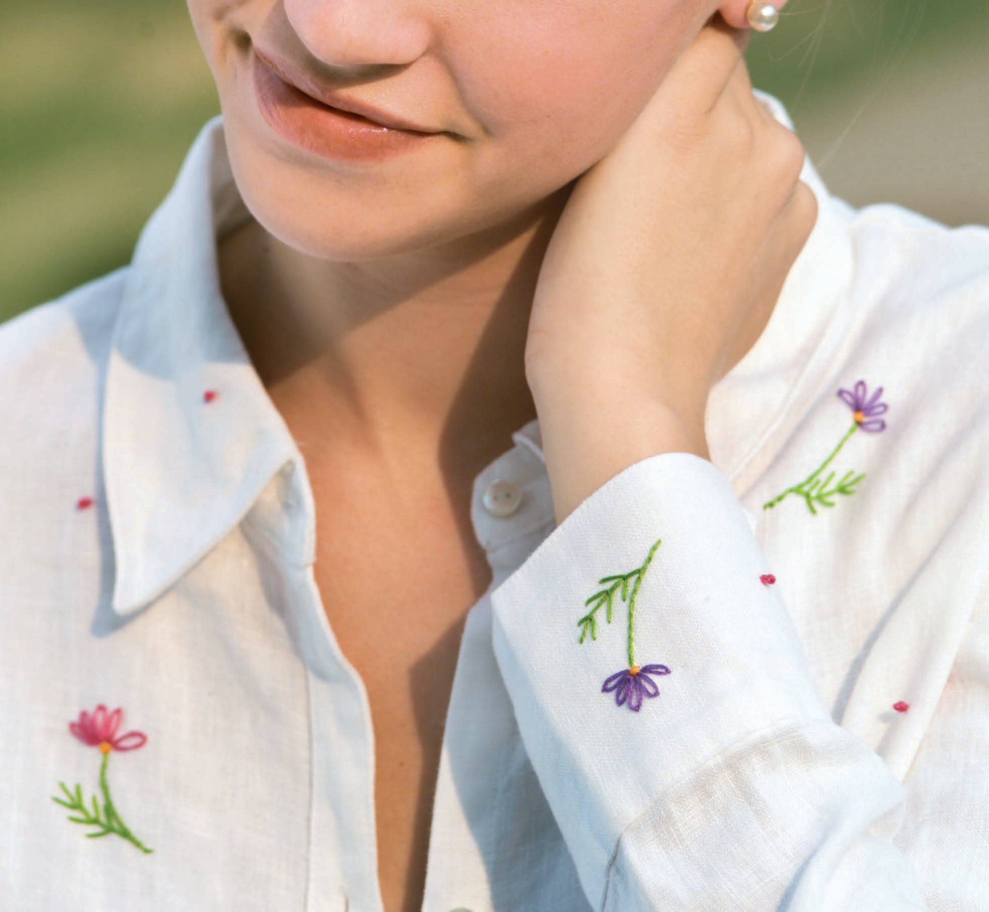 A person models the Sew Simple Embroidery white shirt by Leisure Arts, featuring colorful floral designs ideal for beginner projects. Only their lower face and upper torso are visible, set against a blurred outdoor background.