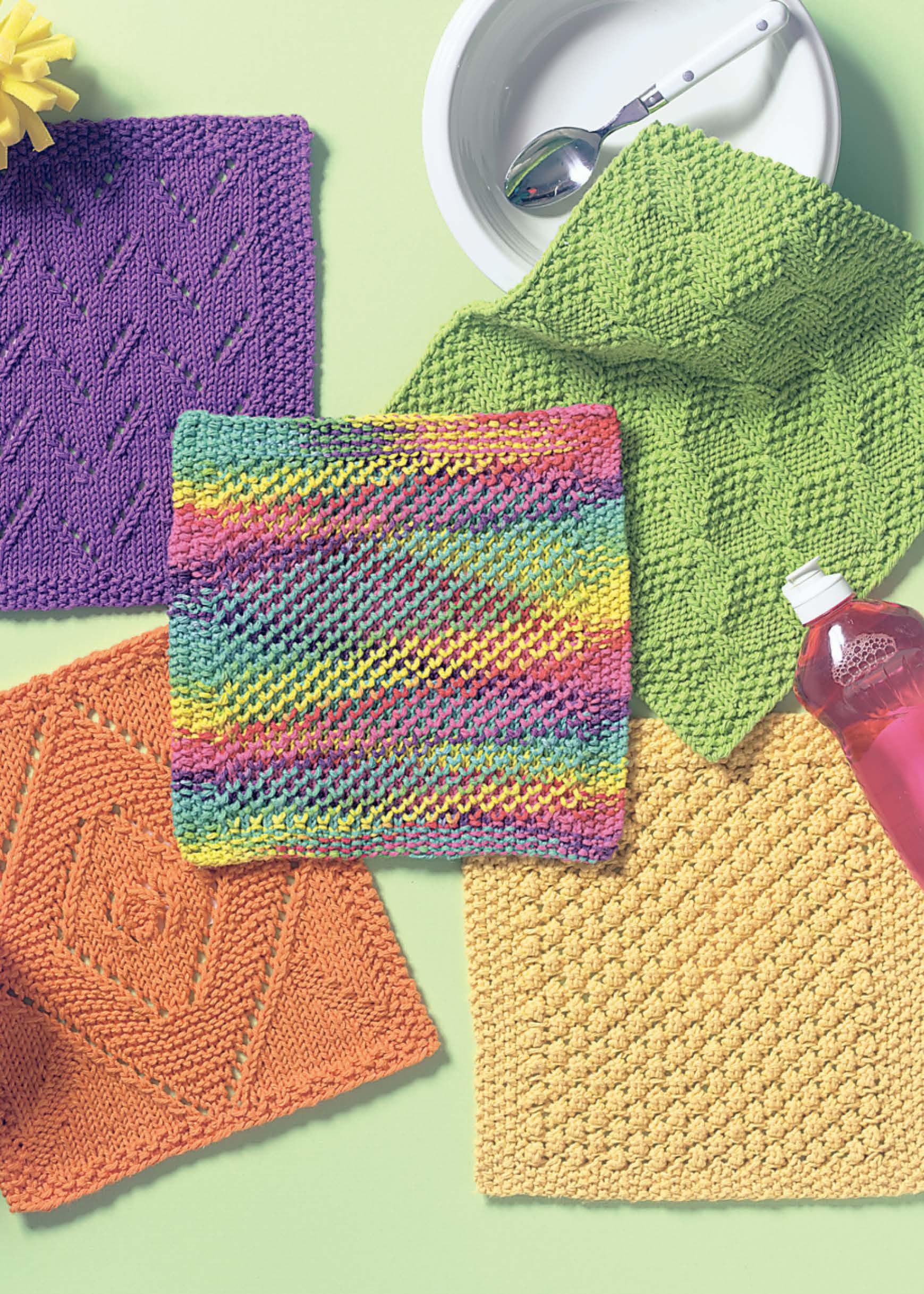 Kitchen Bright Dishcloths by Leisure Arts—five vibrant knit cloths in purple, green, orange, yellow, and multicolor—are arranged around a white bowl with spoon and pink soap bottle, making charming home or gift accessories.
