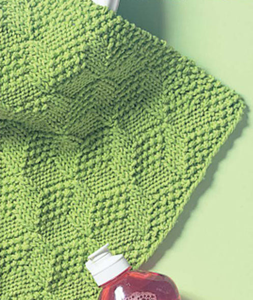 The Kitchen Bright Dishcloth by Leisure Arts, featuring a green diamond knit pattern, rests on a light green surface beside a red dish soap bottle—ideal for home accessories or practicing new stitches.