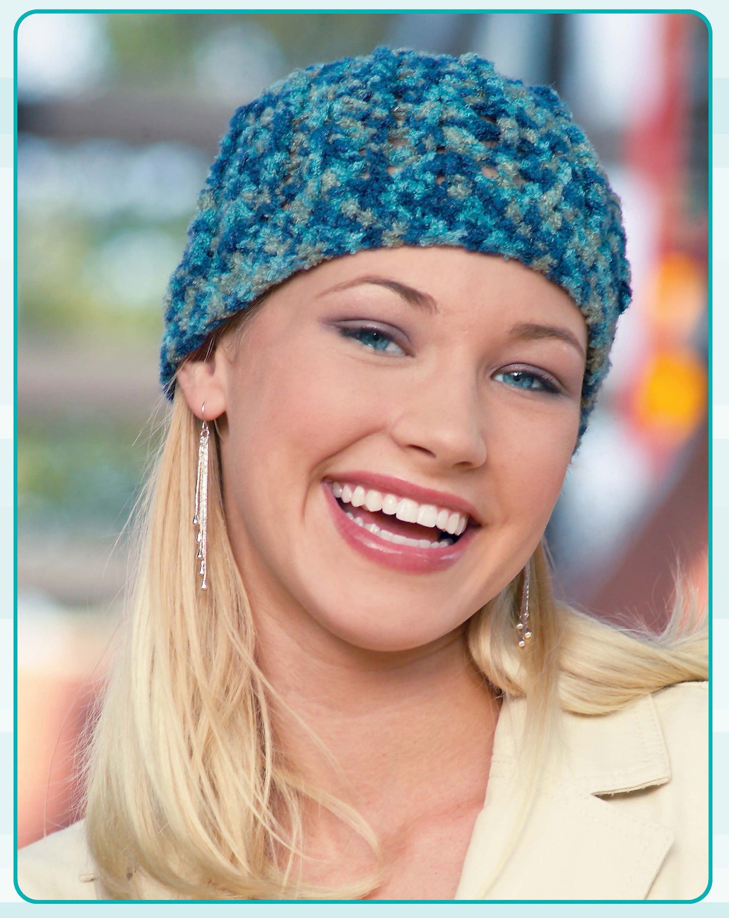 A smiling person with long blonde hair wears a blue and teal hat made from Leisure Arts’ Quick Caps—7 Crochet Designs, plus dangly earrings and a light jacket. The softly blurred background is outdoors.