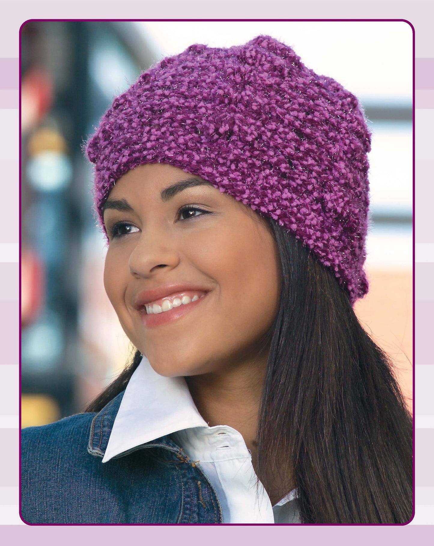 A smiling woman with long dark hair wears a textured purple hat from Leisure Arts' Quick Caps—7 Crochet Designs, styled with a white collared shirt and denim jacket. The softly blurred background shows blue and pink tones.
