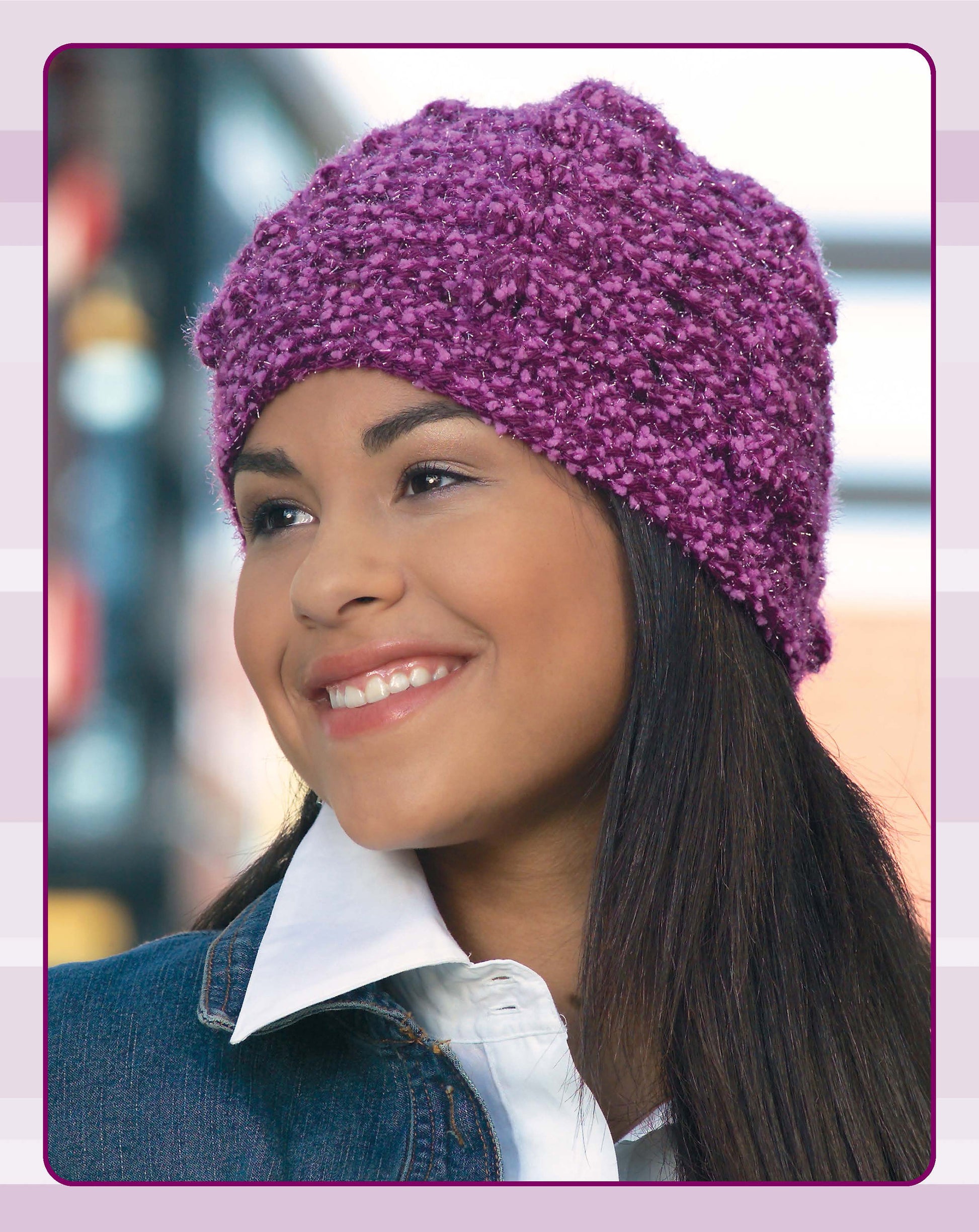 A smiling woman with long dark hair wears a textured purple hat from Leisure Arts' Quick Caps—7 Crochet Designs, styled with a white collared shirt and denim jacket. The softly blurred background shows blue and pink tones.