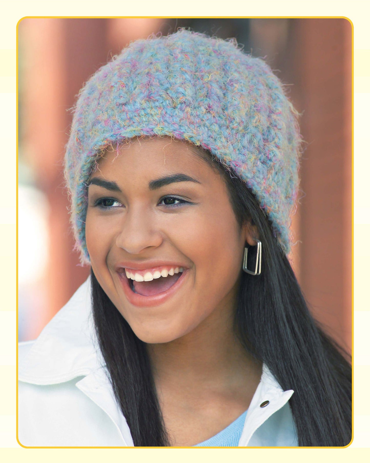 A smiling person with long dark hair wears a pastel textured knit hat from Leisure Arts’ Quick Caps—7 Crochet Designs, along with a white collared jacket and silver hoop earrings, standing outdoors with a blurred background.