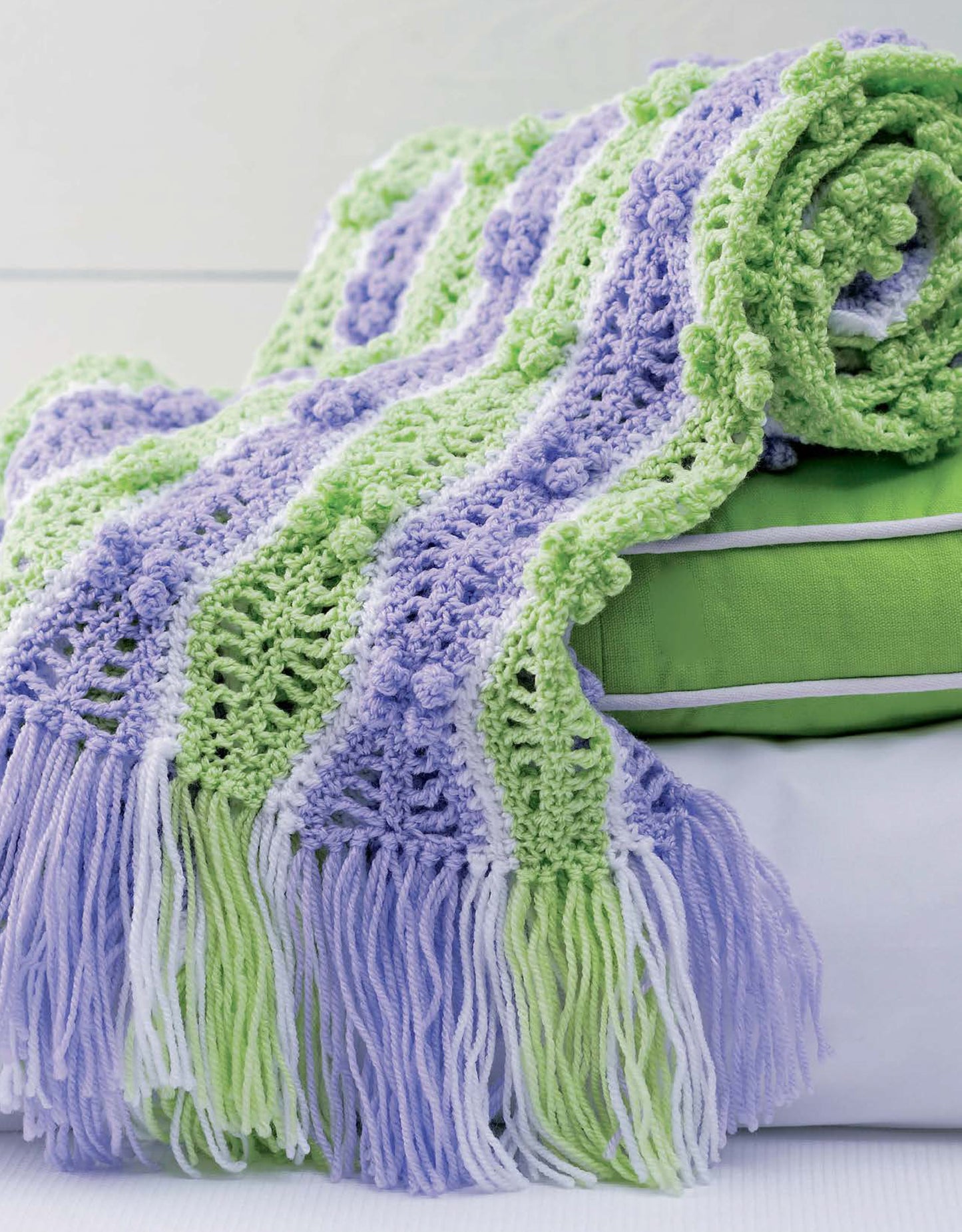 A folded green and white pillow with a crocheted Leisure Arts "Baby Bubble Wraps—6 Afghans" blanket draped over it. The sport-weight blanket has pastel green, lavender, and white wavy stripes with long matching fringes.