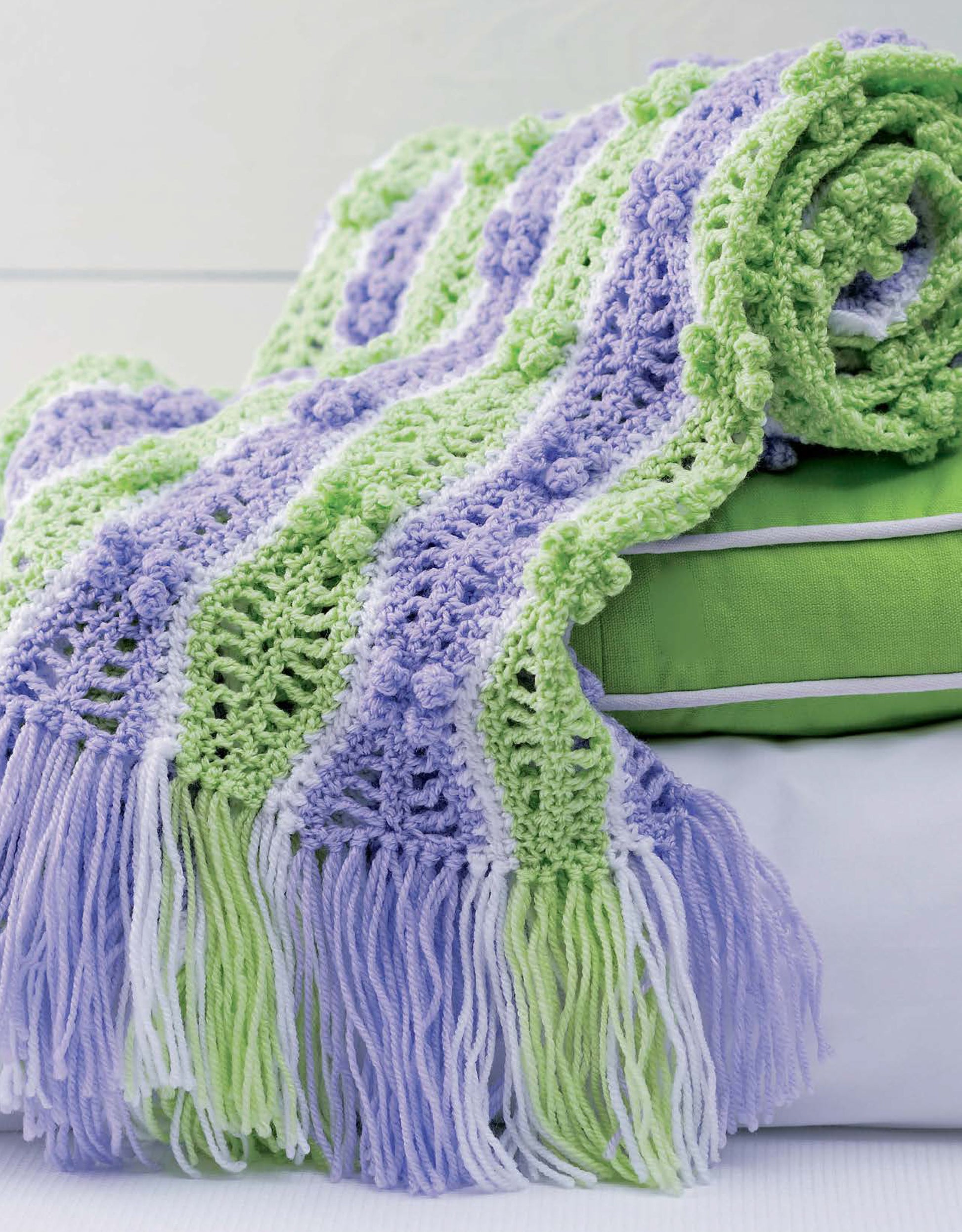 A folded green and white pillow with a crocheted Leisure Arts "Baby Bubble Wraps—6 Afghans" blanket draped over it. The sport-weight blanket has pastel green, lavender, and white wavy stripes with long matching fringes.