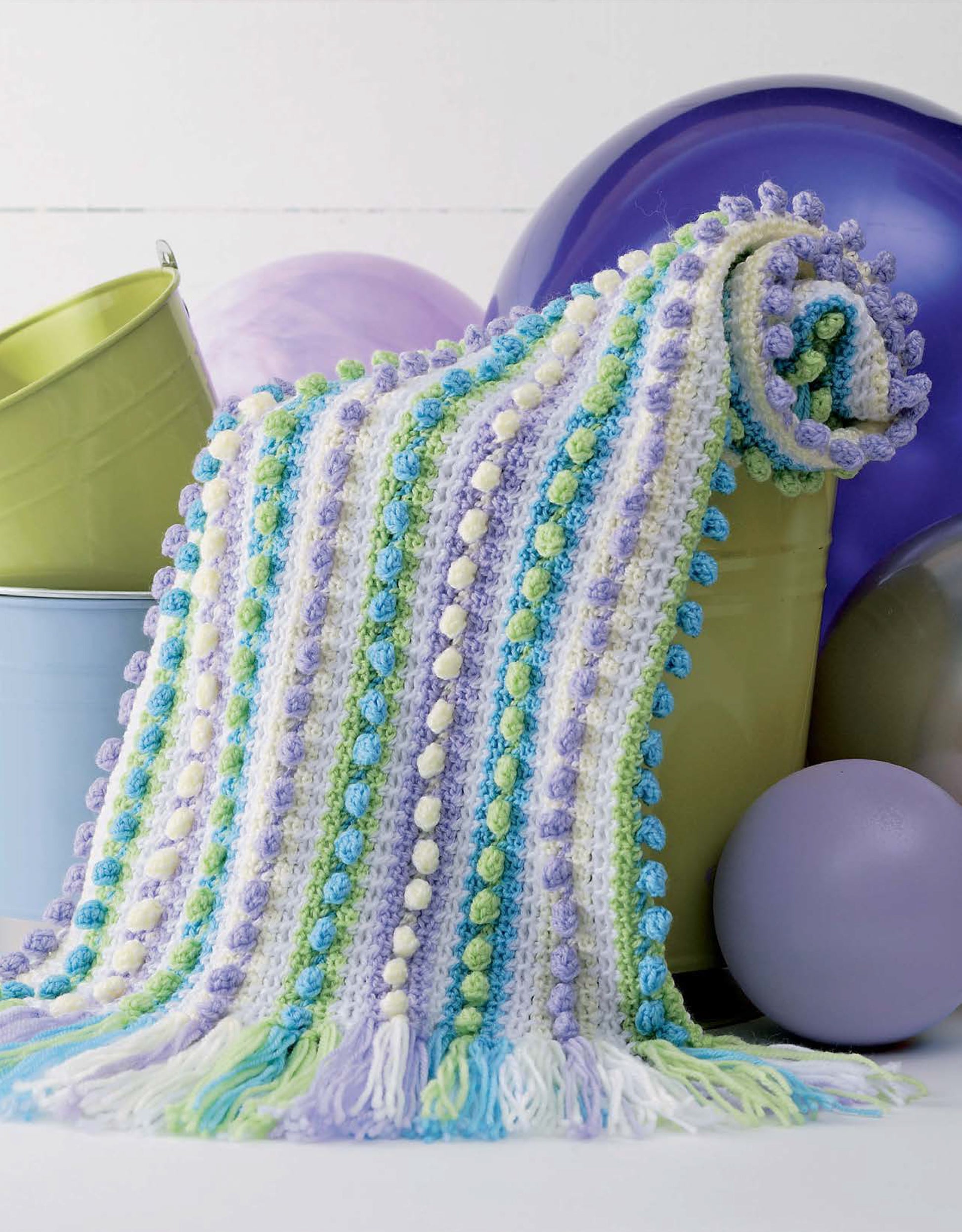 The "Baby Bubble Wraps—6 Afghans" by Leisure Arts, a pastel crocheted blanket with bobble and fringe details, is draped over a green bucket amid blue, green, and purple balloons, with another bucket in the background.