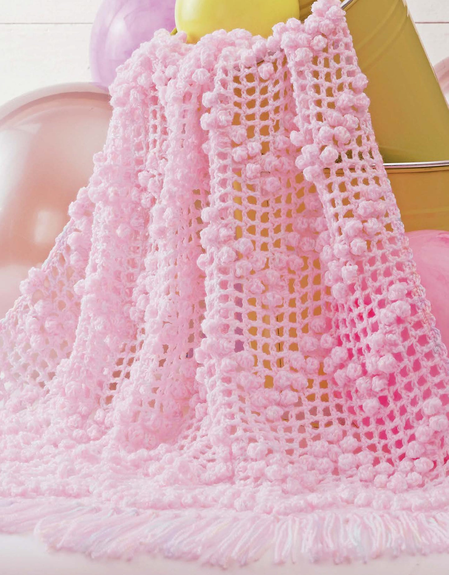 The Baby Bubble Wraps—6 Afghans by Leisure Arts features a soft, light pink crochet afghan with a textured bobble pattern and fringe, draped over a yellow bucket amid pastel balloons.