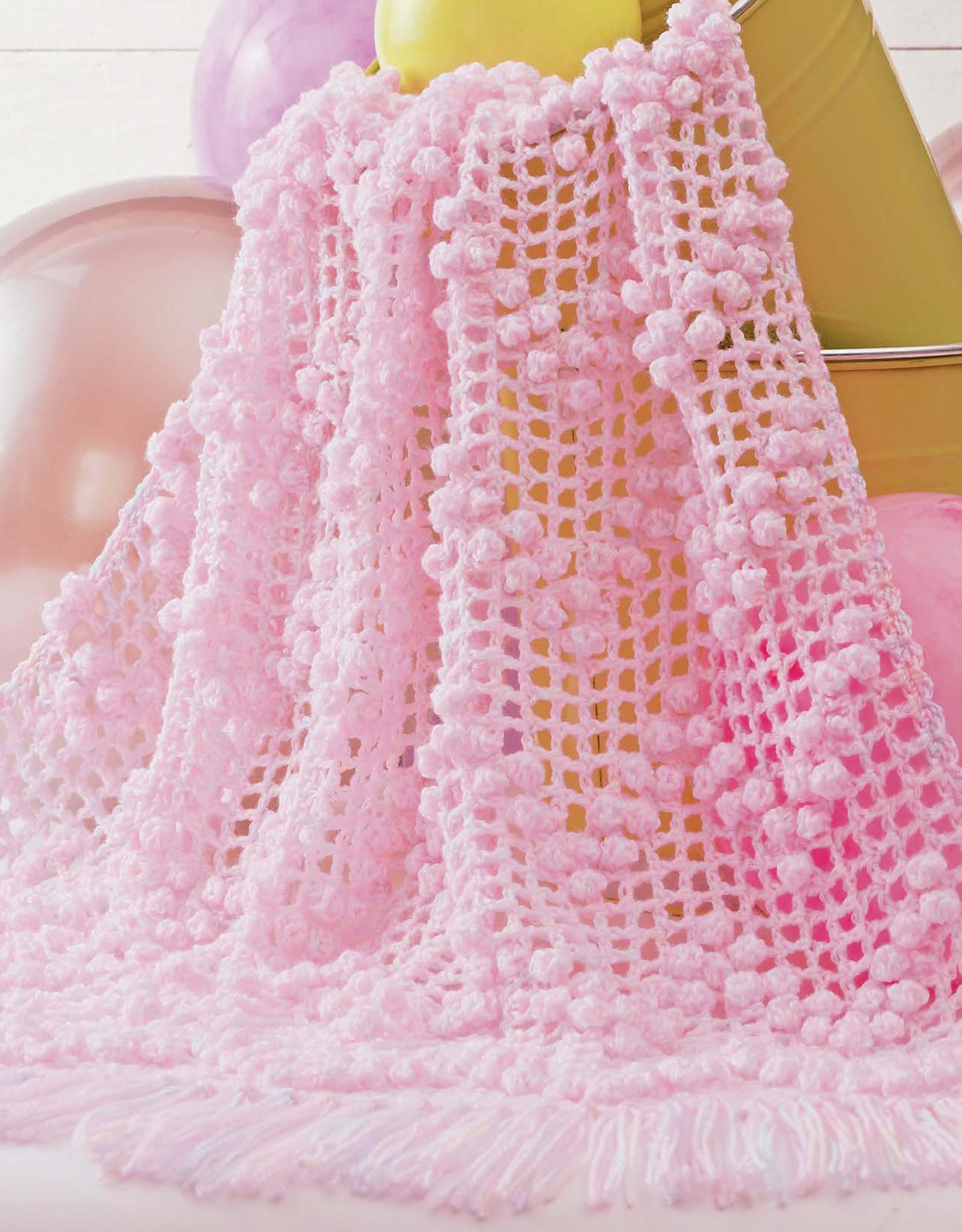The Baby Bubble Wraps—6 Afghans by Leisure Arts features a soft, light pink crochet afghan with a textured bobble pattern and fringe, draped over a yellow bucket amid pastel balloons.