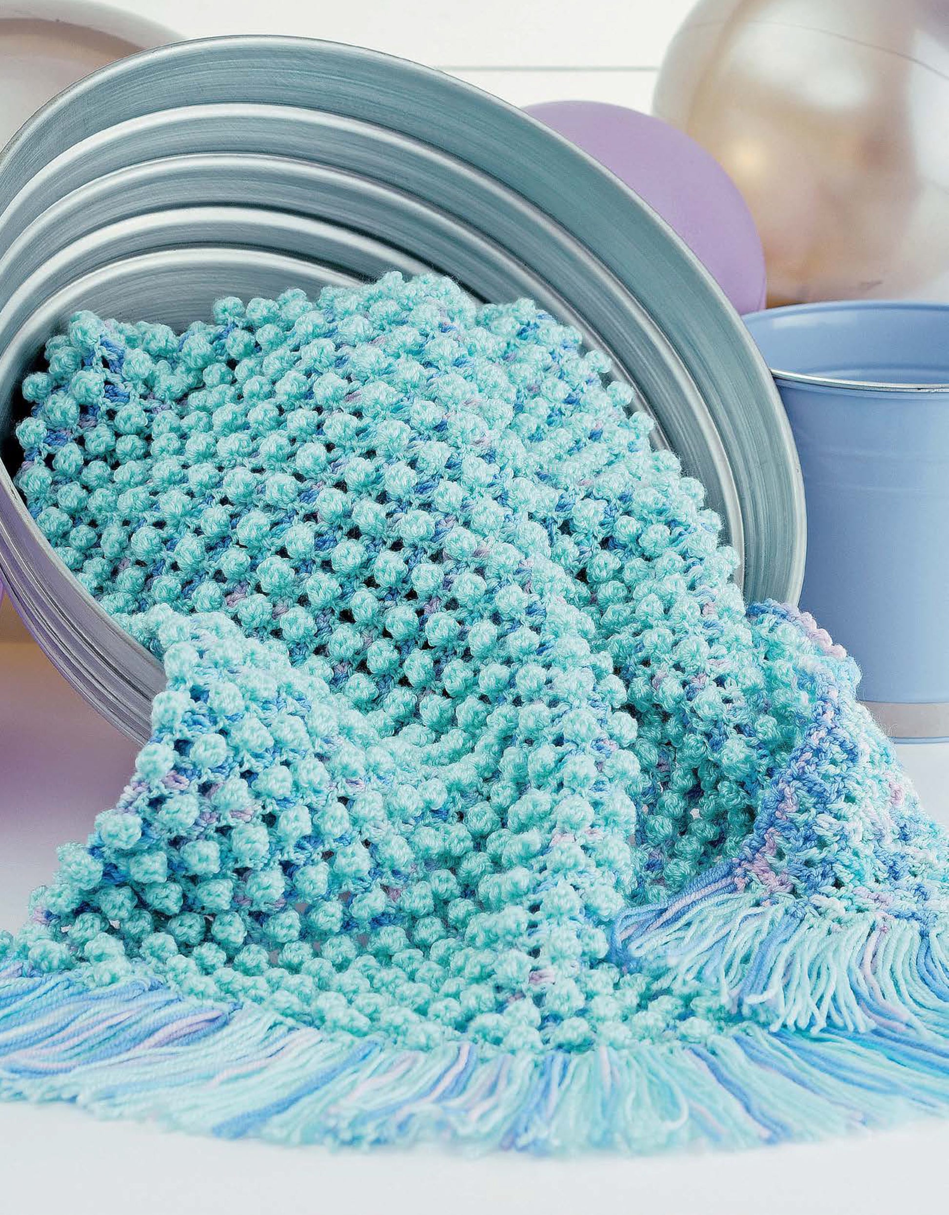 The Leisure Arts Baby Bubble Wraps—6 Afghans, shown in aqua blue and lavender with cluster and bobble stitches plus fringe, is draped over metal buckets with pastel balloons and a light blue cup in the background.