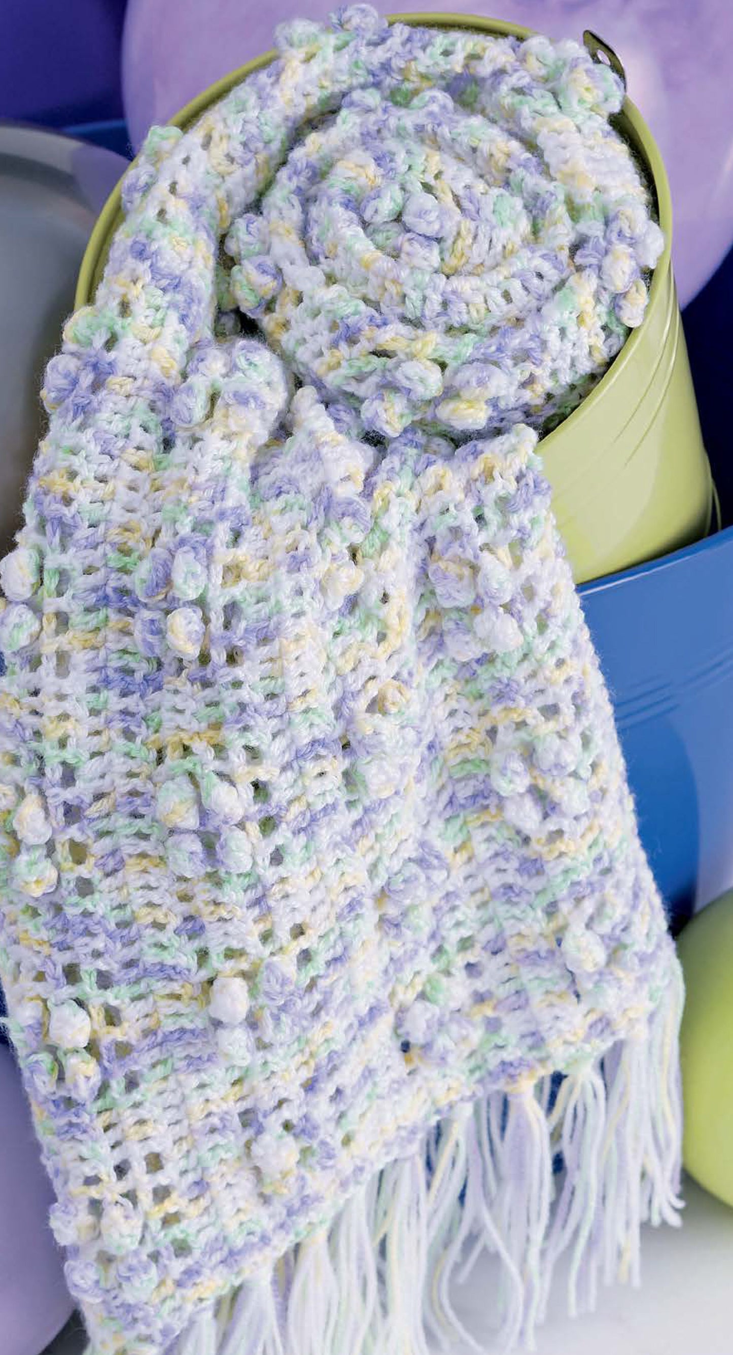 A pastel crochet scarf from Baby Bubble Wraps—6 Afghans by Leisure Arts, made with variegated sport yarn in soft purple, yellow, blue, and white, featuring cluster and bobble stitches plus fringe, is draped over a green container.