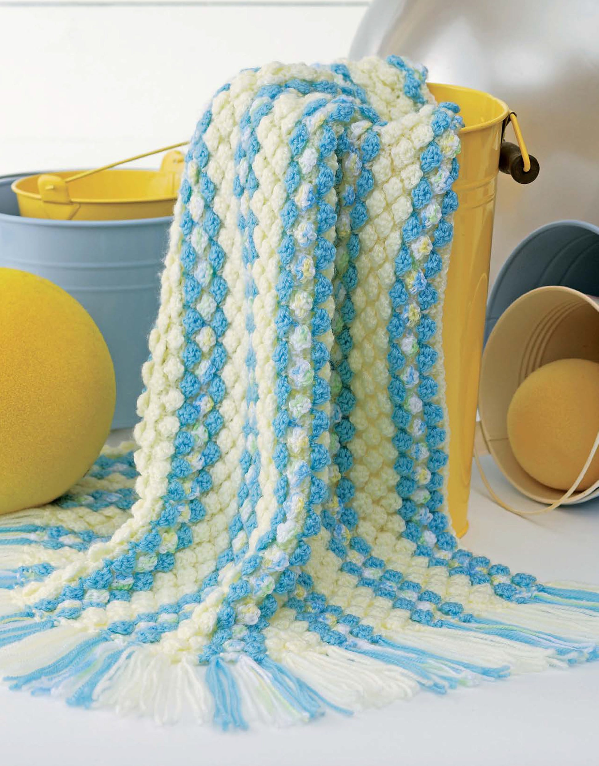 The Leisure Arts Baby Bubble Wraps—6 Afghans features a crocheted sport weight blanket with blue, white, and yellow stripes on a yellow bucket, set before a blue bucket, yellow sponge ball, and beige and blue sand pails.