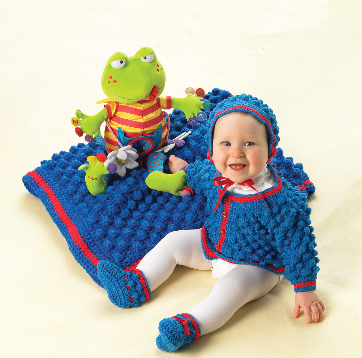 A smiling baby wearing a crocheted ensemble from Leisure Arts' Bright Layettes to Crochet sits on a matching blanket beside a green frog plush in a striped shirt, looking up and reaching toward the camera.